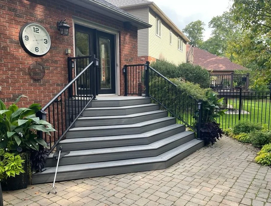 A set of modern, gray wooden steps with black metal railings leading to a door on a brick house, with a large round clock on the wall, surrounded by green plants and a paved walkway, and a fenced yard with trees in the background.