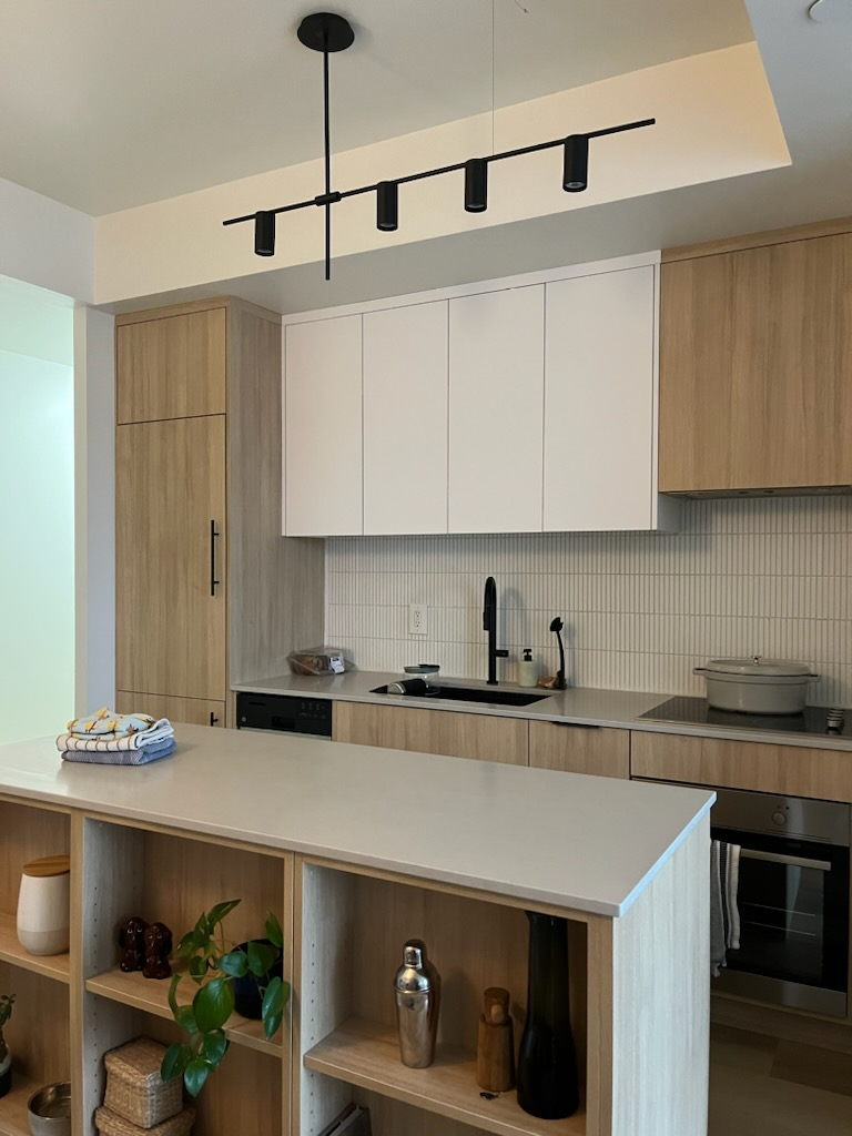 Modern kitchen with wood cabinets, white upper cabinets, black faucet, built-in oven, and decorative items on a light wood shelf.