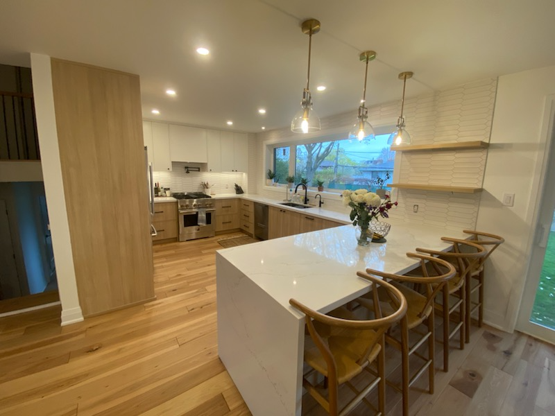 Modern kitchen with wooden floors, white and wood cabinets, a large white island with a flower vase, four wooden bar stools, and pendant lighting over the island. Large window with a view of trees outside.