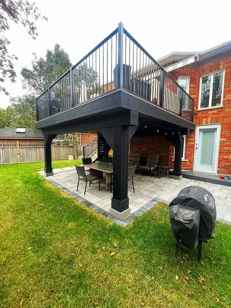 Backyard patio with a covered area and a second-story deck with black railing attached to a brick house.