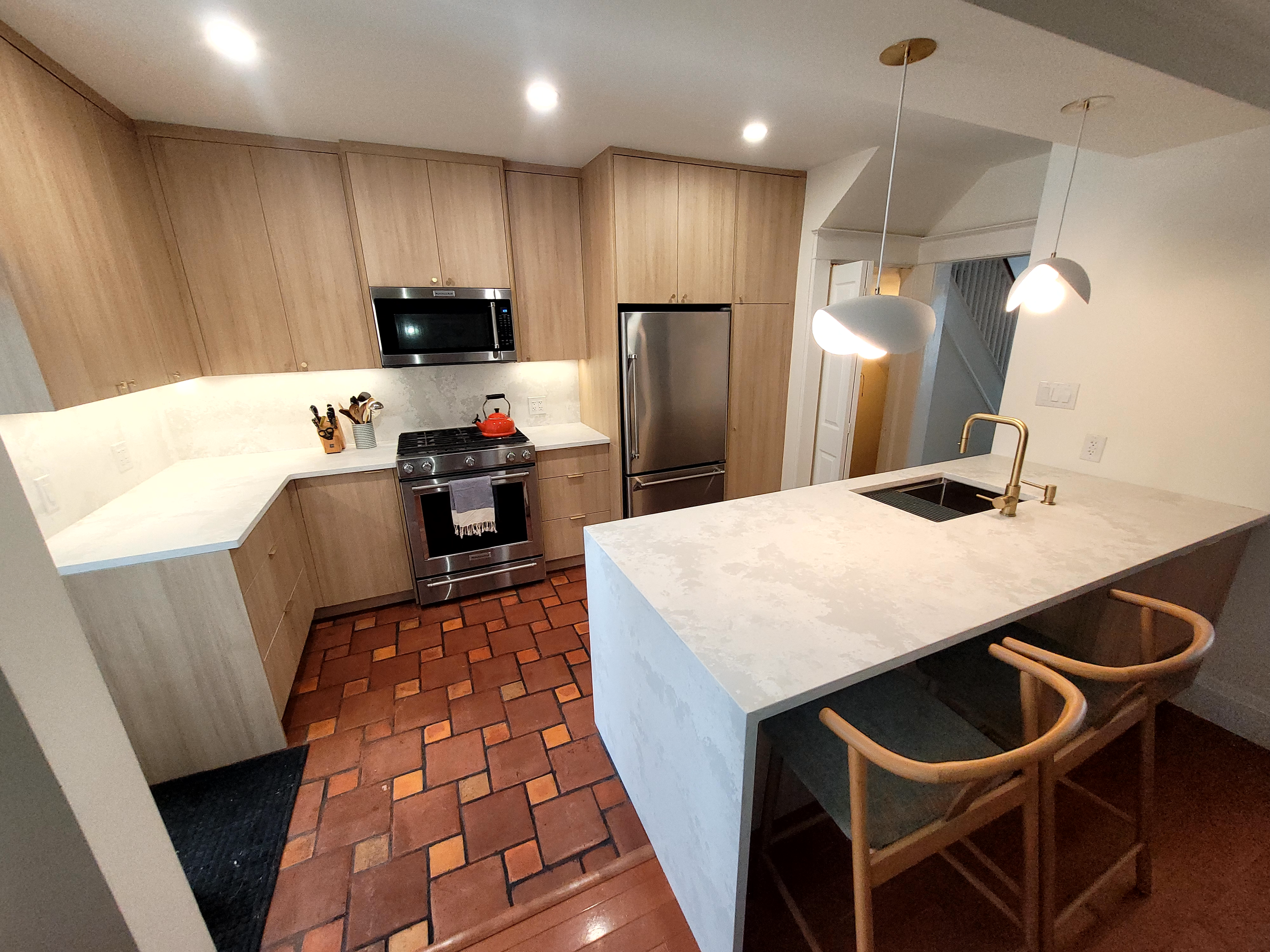 Modern kitchen with light wood cabinets, stainless steel appliances, white countertops, hanging pendant lights, tiled brick floor, and two wooden bar stools at a peninsula.