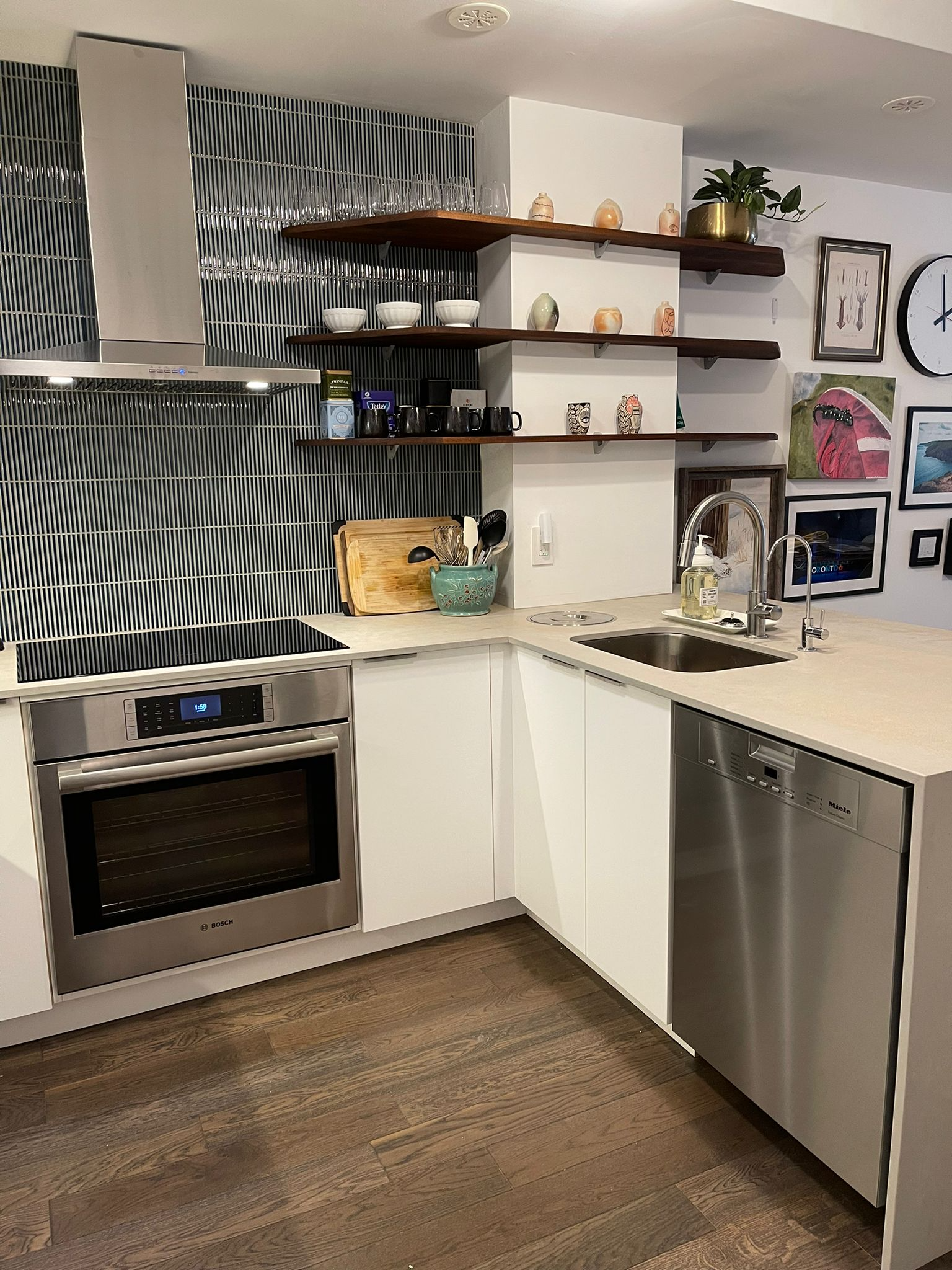 Modern kitchen with white cabinetry, stainless steel oven and dishwasher, black tile backsplash, open wooden shelves with dishes, glasses, and decorative items, stainless steel sink with soap dispenser, and various wall art and clocks in the backgrou