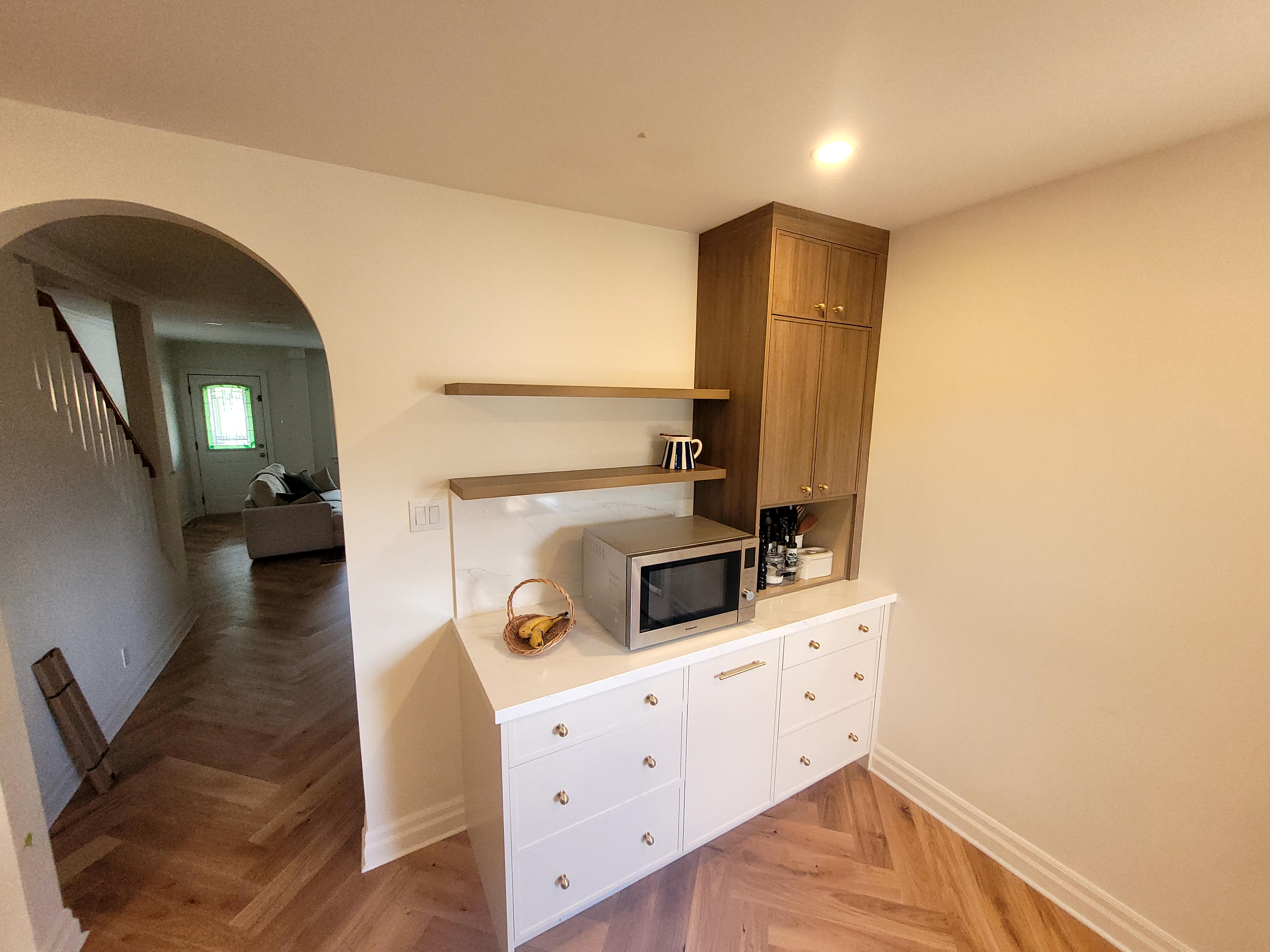 Kitchen corner with white cabinet, microwave, wooden gift basket, open wooden shelving, and upper cabinetry, hardwood flooring, and beige walls.