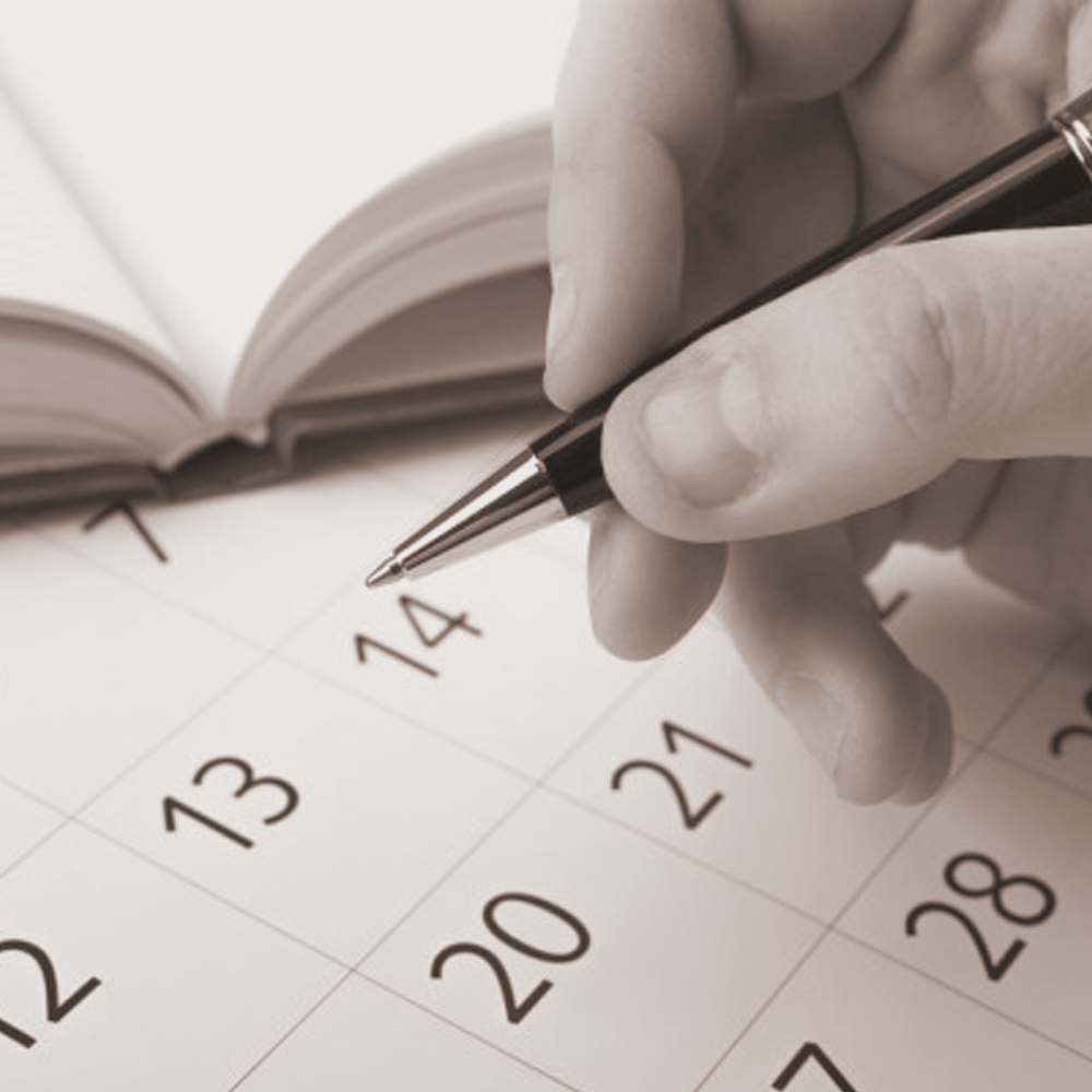 Close-up of a person writing on a calendar with a pen, with an open book in the background.