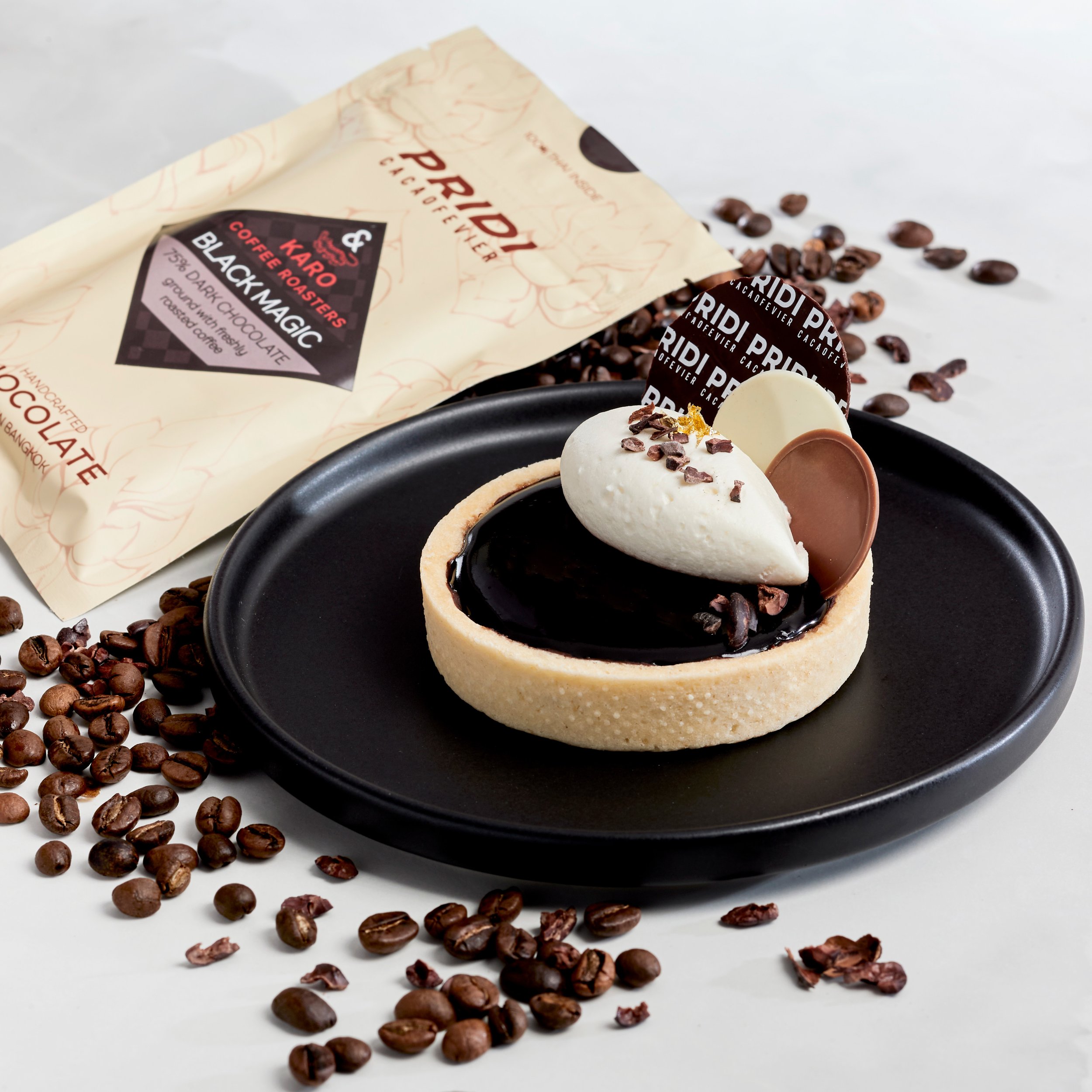 Chocolate tart topped with cream and chocolate decorations on a black plate, surrounded by coffee beans. Packaged chocolate in the background.