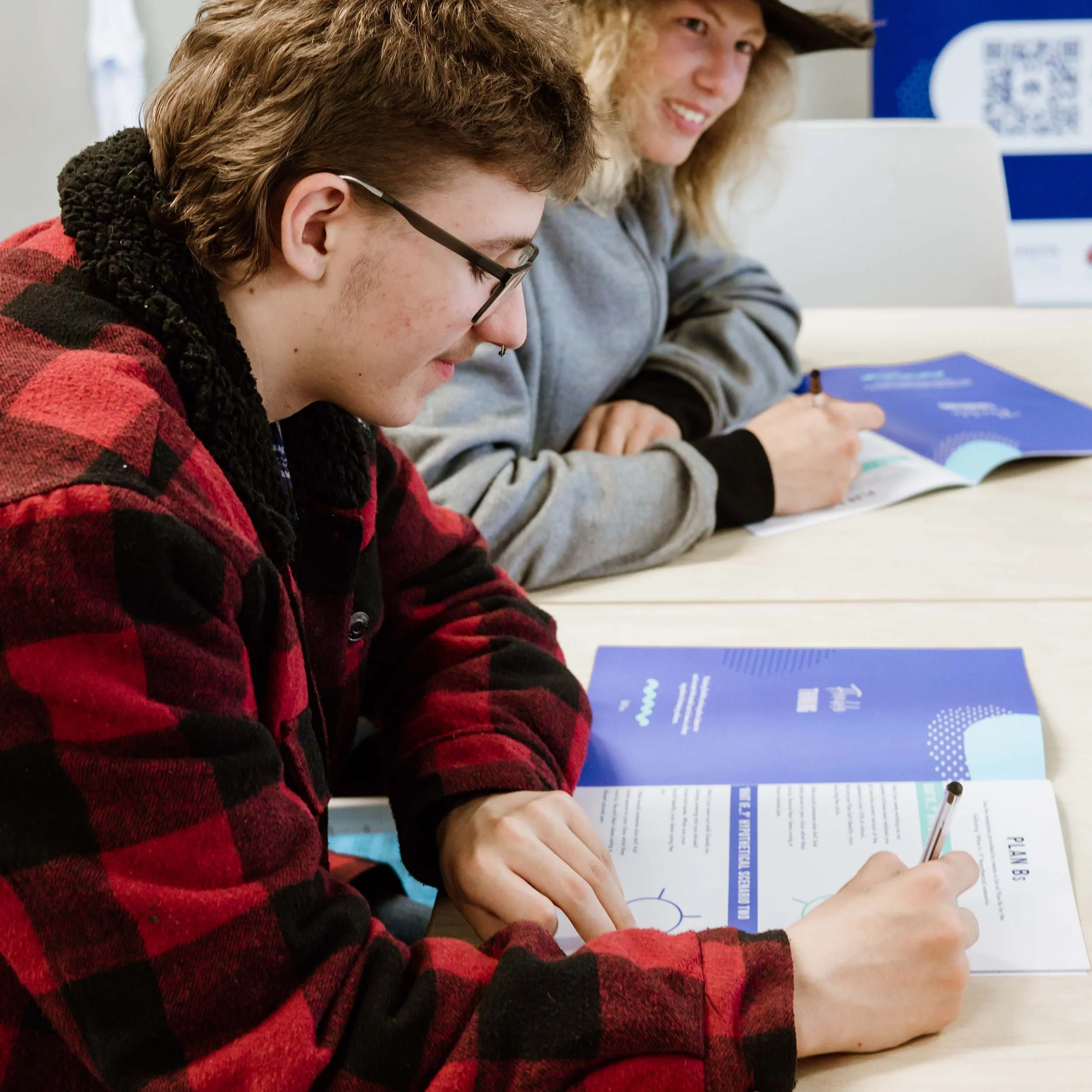 2 teenagers sit at a school desk, working through their RAMS student workbook.