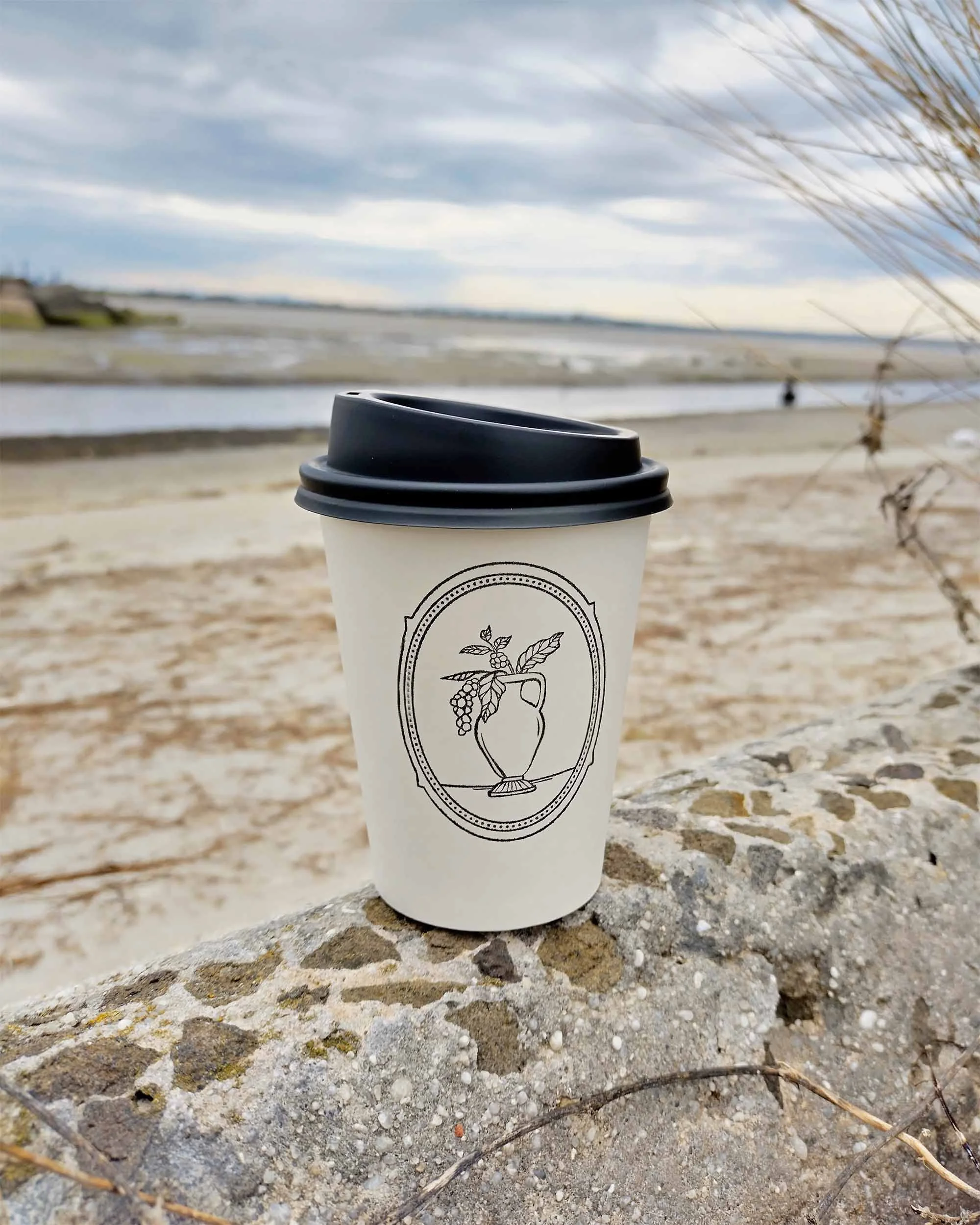 A white takeaway coffee cup, stamped with the Baci's logo, sits on a stone wall facing the ocean.