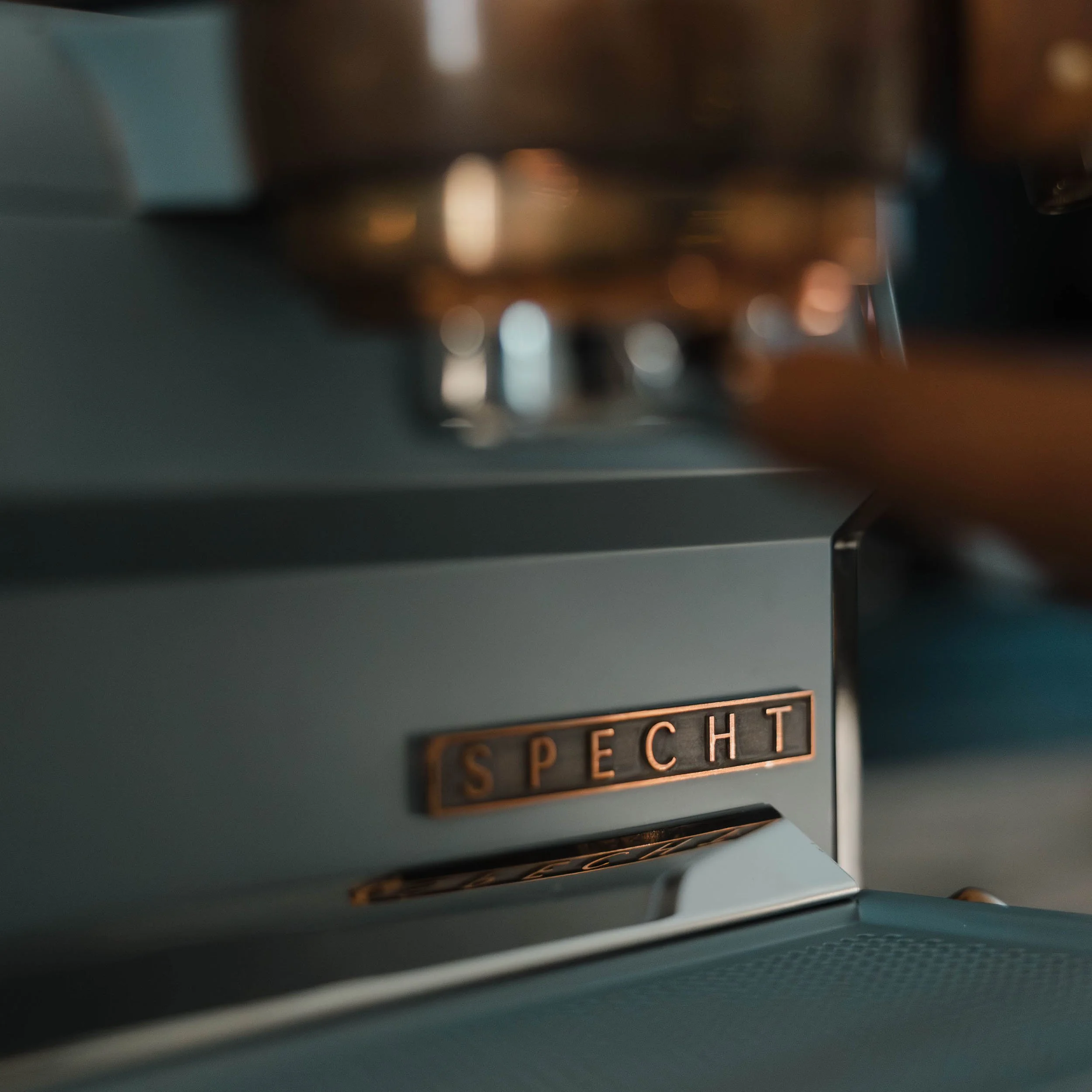 A brass Specht logo on a teel Slayer espresso machine.