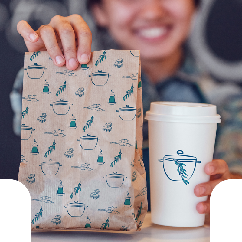 A female barista holds a Kazan branded takeaway bag and coffee cup.