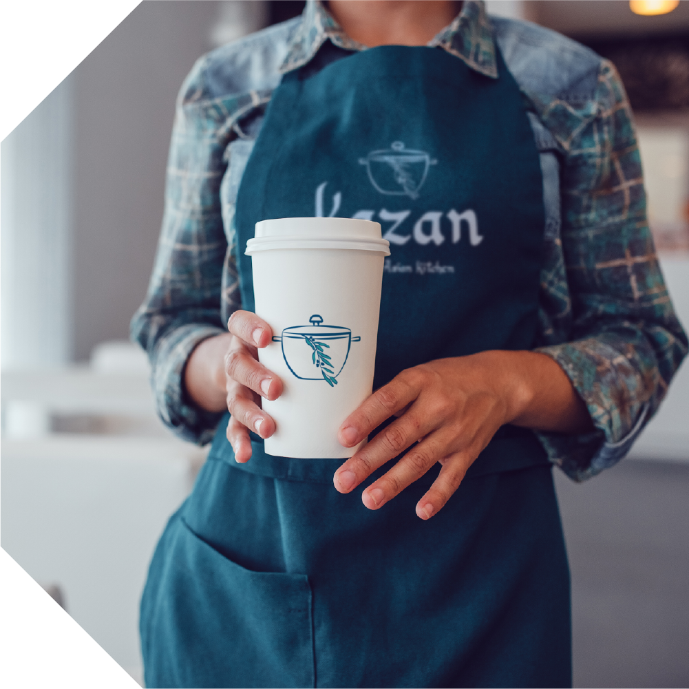 A female barista is holding a white takeaway coffee cup with the Kazan logo stamped on it, and is wearing a blue, Kazan-branded apron.