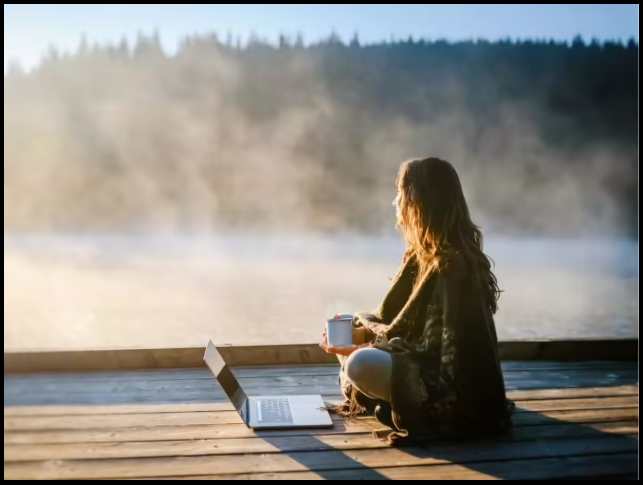 A woman sitting on a wooden dock by a lake, holding a mug, with a laptop open nearby. The scene is illuminated by warm sunrise light, with mist over the water and trees in the background.