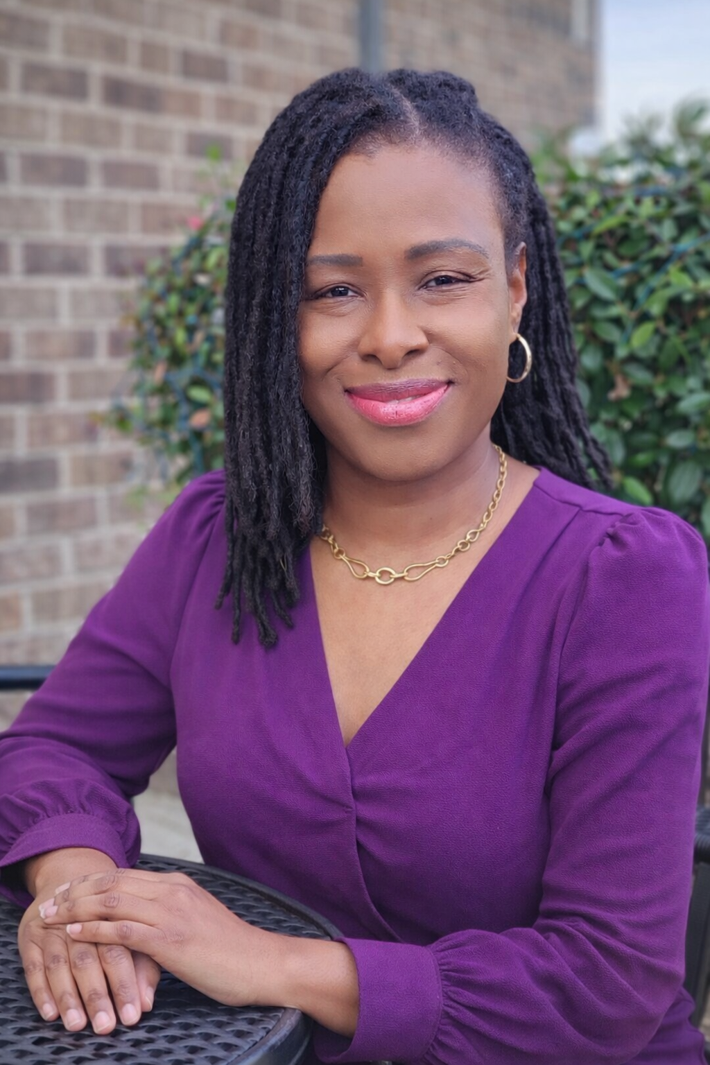 A woman with shoulder-length dreadlocks, wearing a purple top and gold jewelry, sitting at an outdoor table with a brick wall and green shrubbery in the background.