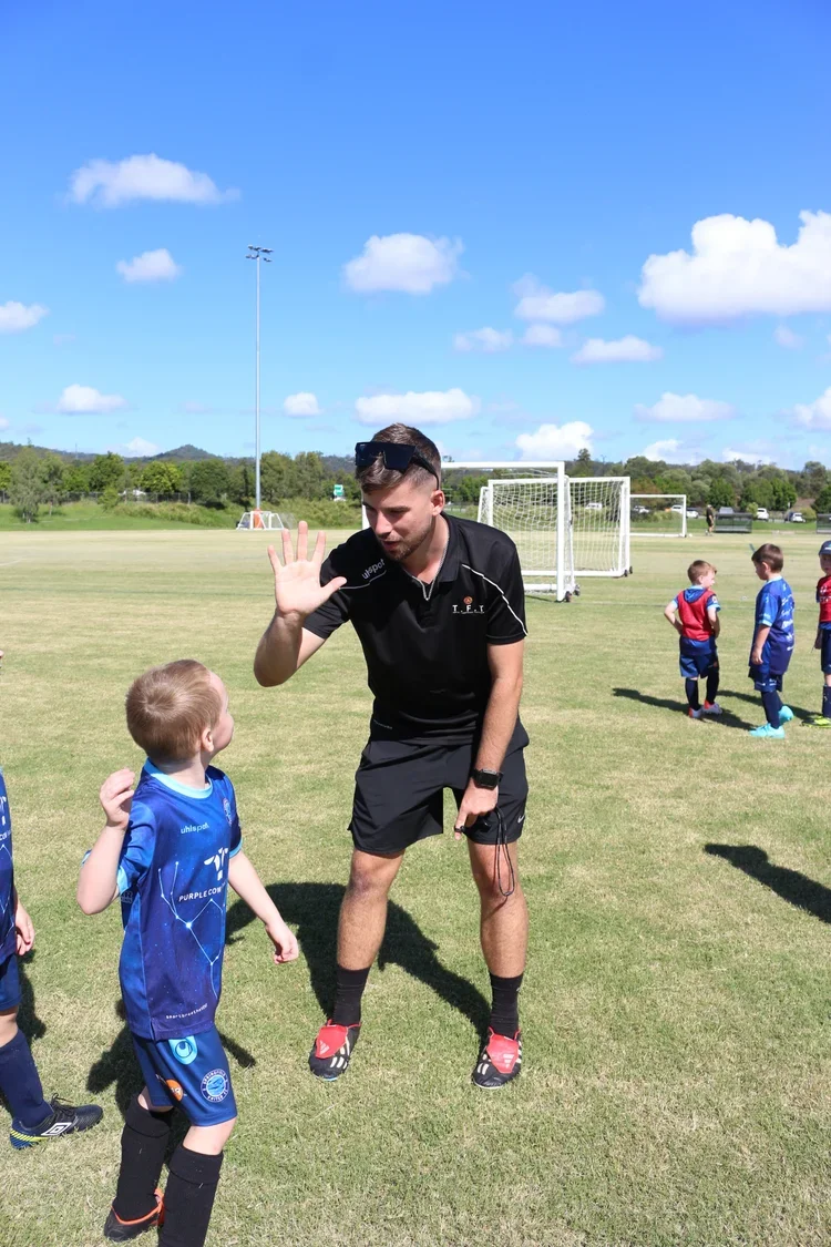 Male coach wearing black TFT shirt holding his hand up to high-five a young boy in blue