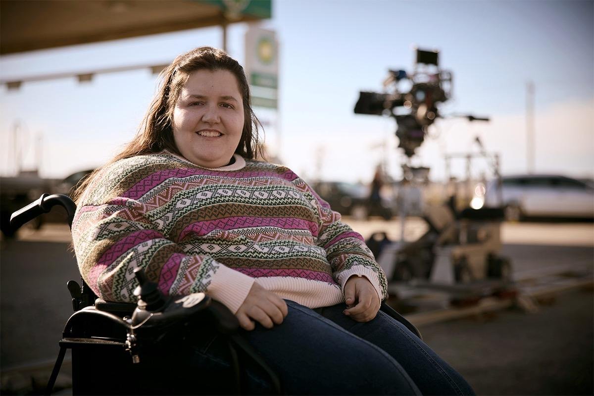 A smiling woman wearing a colourful patterned jumper sits in a powered wheelchair outdoors, with a film camera and petrol station blurred in the background.