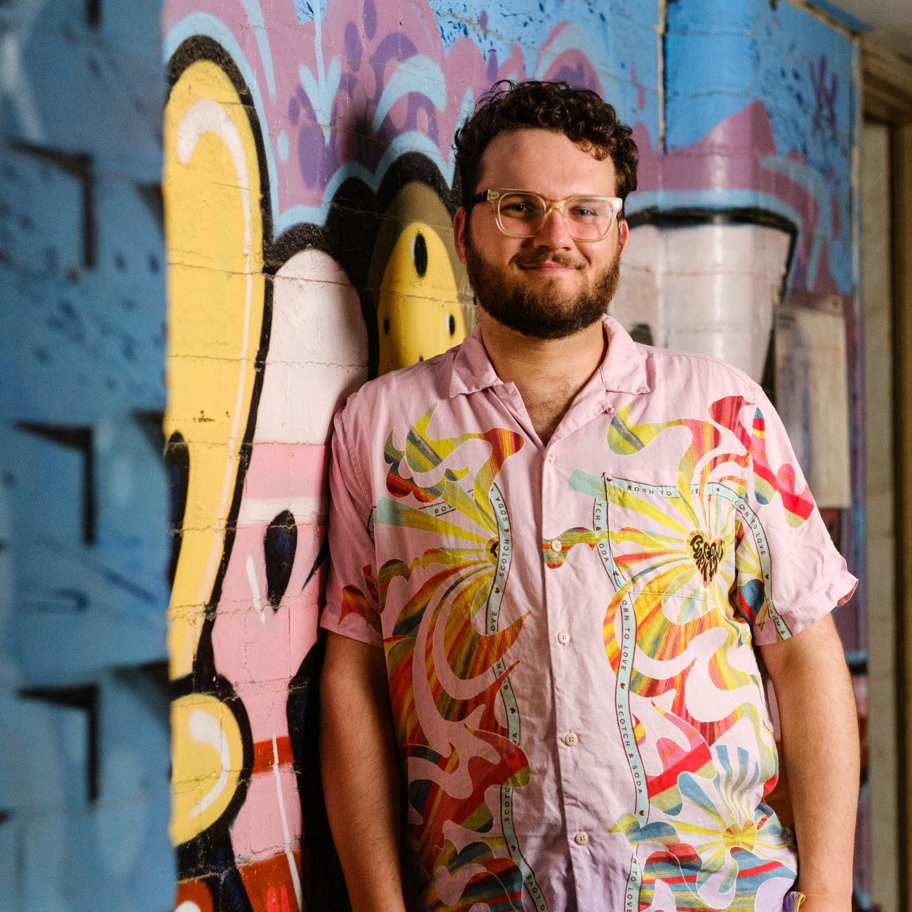 A smiling bearded man wearing glasses and a colourful pink patterned shirt leans against a brightly painted mural in an indoor urban space.