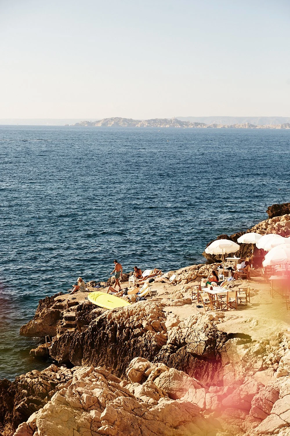 People relaxing on rocky coast under umbrellas with ocean on the background and distant land across the water.