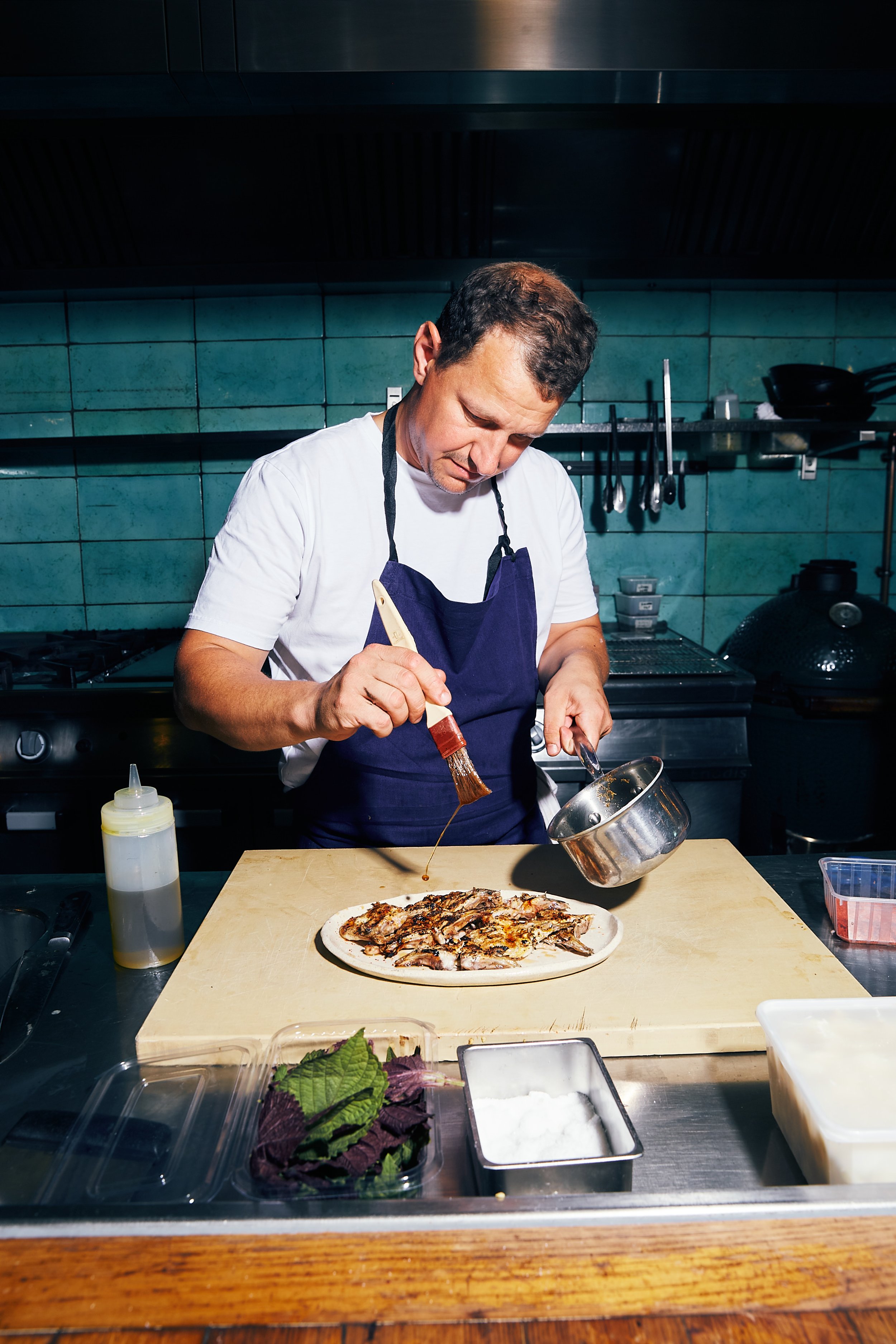 Harry Cummins in the kitchen at La Mercerie, shot by Lizzie Mayson for Table Magazine, pouring sauce on grilled fish on a wooden cutting board.