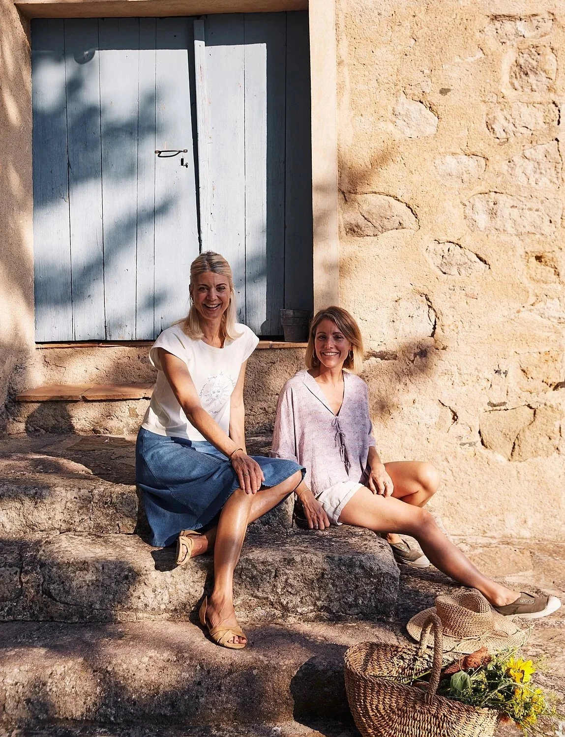 Frankie Unsworth and Jeany Cronk sitting on stone steps outside a rustic building with a blue wooden door, smiling and enjoying the sunlight, with a basket of flowers on the ground nearby.