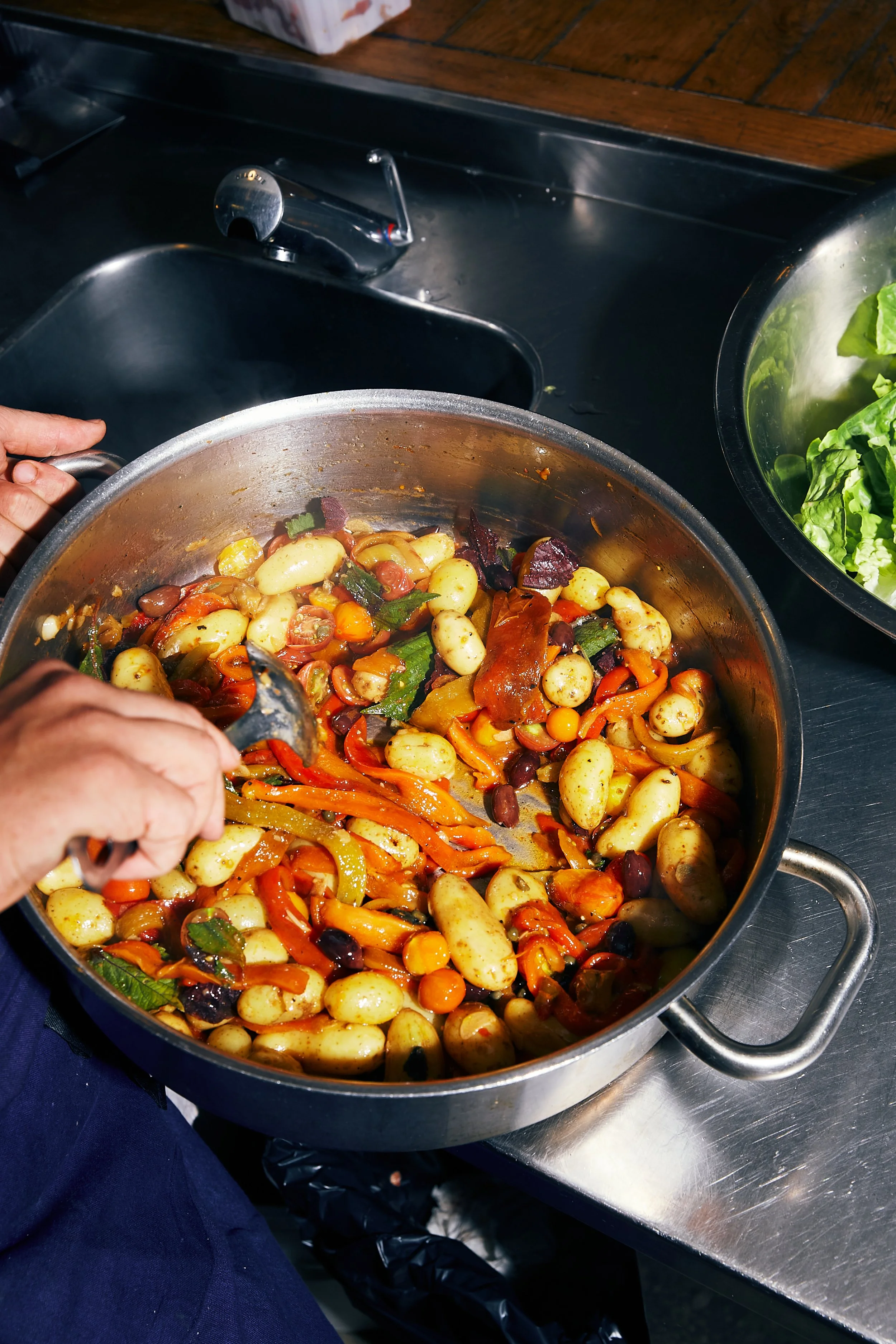 Person scooping mixed cooked vegetables, including gnocchi, bell peppers, and olives, from a large metal pot in a kitchen.