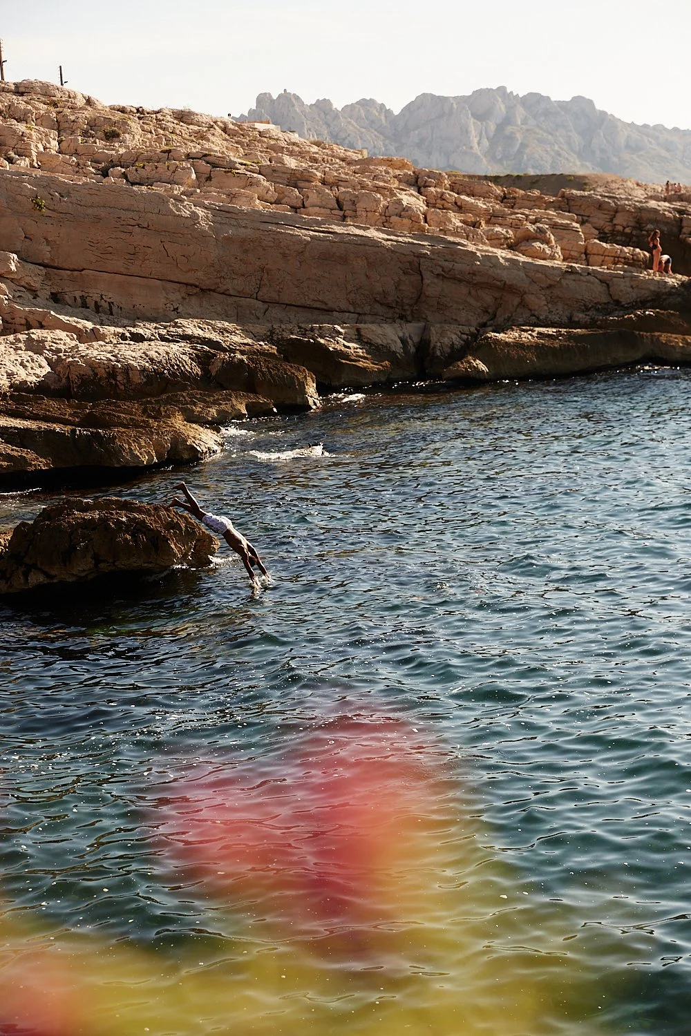 A person diving into the water from a rocky shoreline during daytime, with mountains in the background.
