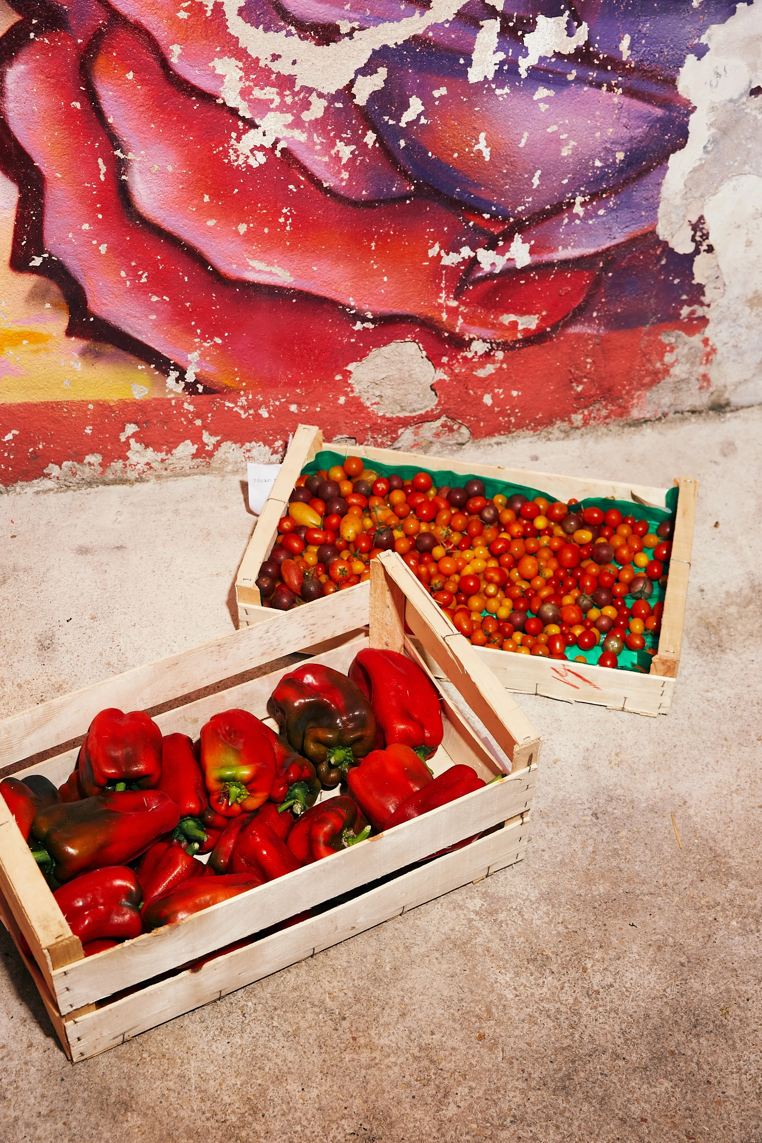 Wooden crates filled with red and green bell peppers and cherry tomatoes, placed on a concrete ground in front of a colorful, weathered mural wall.