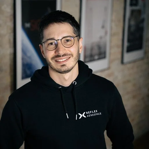 An image of Sam Bender, head of Marketing and Growth at Reflex Aerospace, wearing glasses and a short beard and a black hoodie with the Reflex Aerospace logo standing indoors against a brick wall with framed pictures.