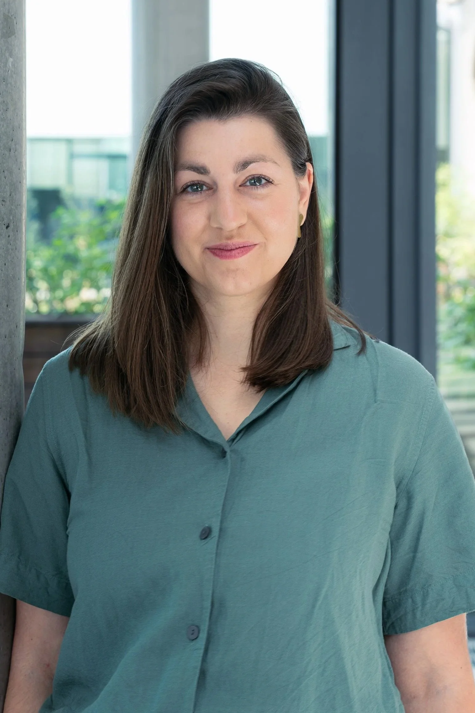 A photo of the VP People and Culture from Reflex Aerospace with long brown hair, wearing a teal button-up shirt, standing indoors near a concrete pillar and large windows.