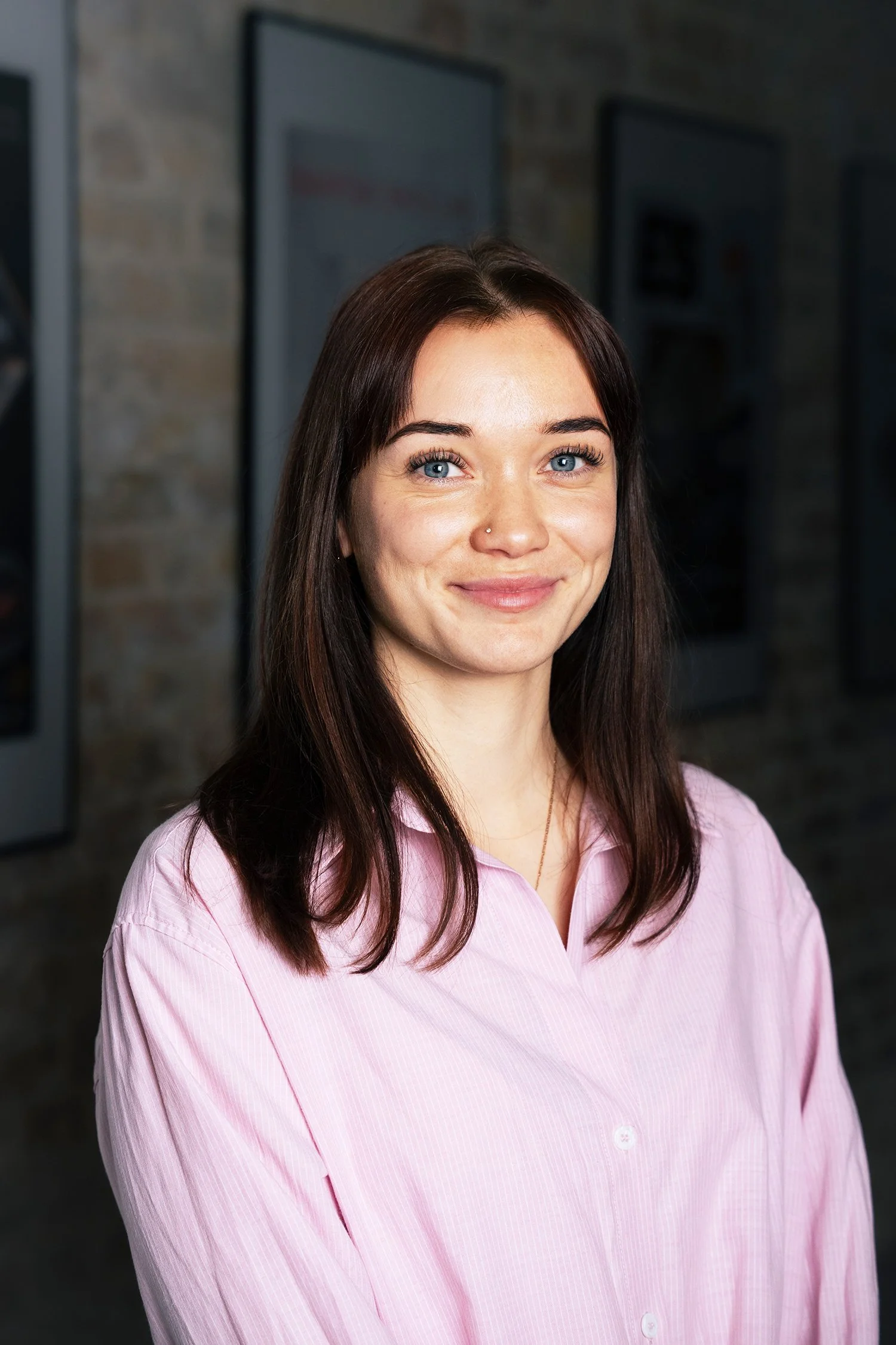 Junior HR Specialist, Paula Schoene, a young woman with shoulder-length dark brown hair, blue eyes, and a nose piercing, smiling at the camera while wearing a light pink shirt, standing indoors against a background of framed pictures on a brick wall.