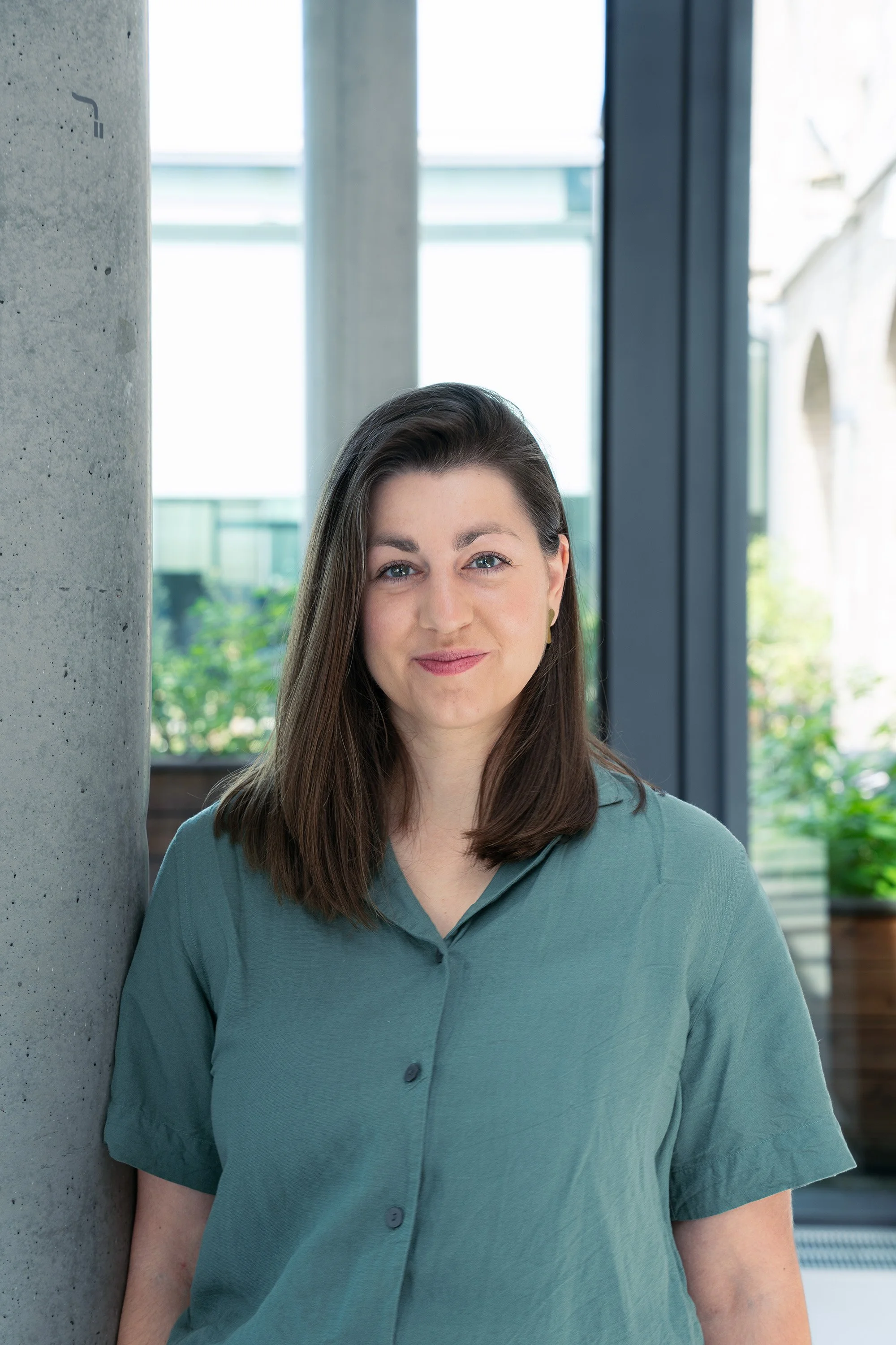 A photo of the VP People and Culture from Reflex Aerospace with long brown hair, wearing a teal button-up shirt, standing indoors near a concrete pillar and large windows.