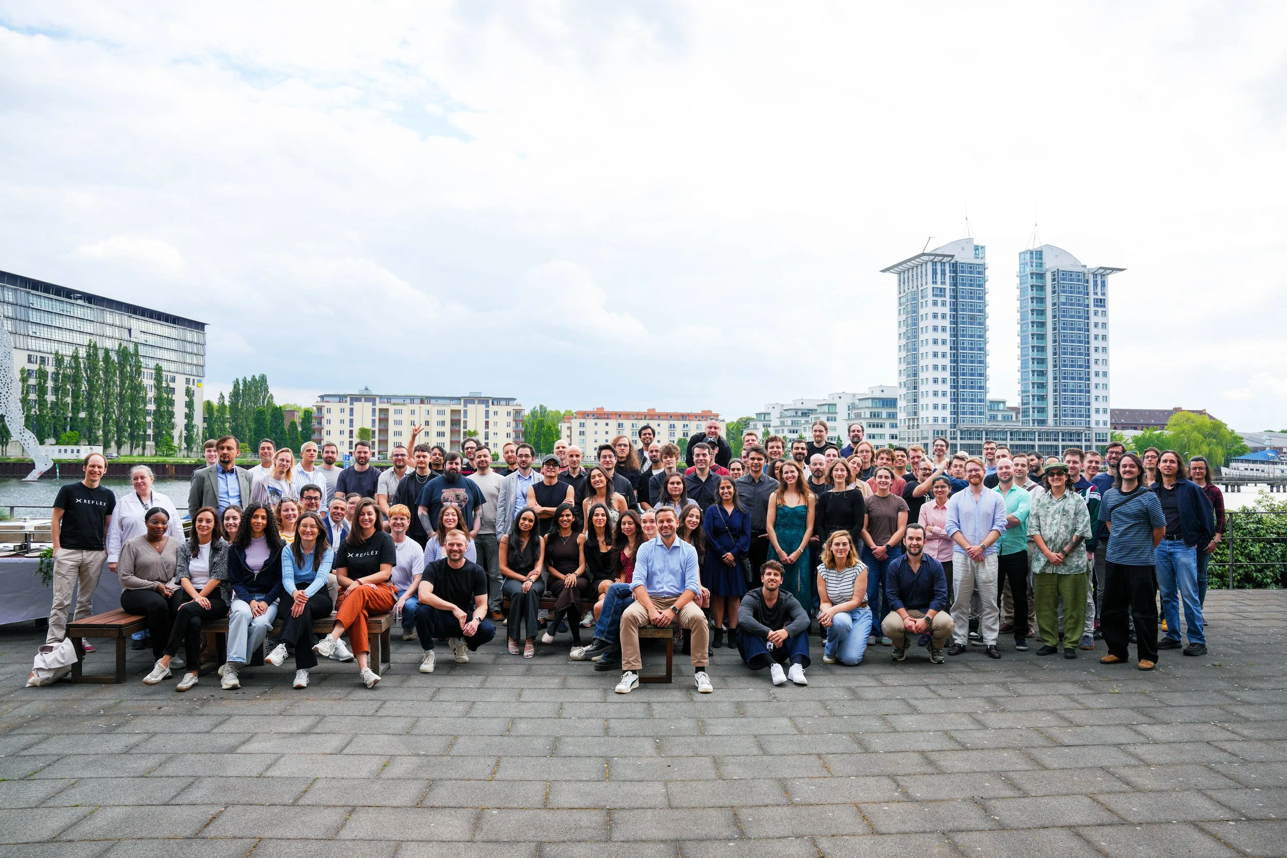 Group of employees from Reflex Aerospace people posed outdoors near a river in Berlin with modern buildings and trees in the background.