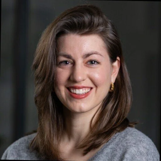 A woman with brown hair smiling, wearing a gray top and gold earrings.