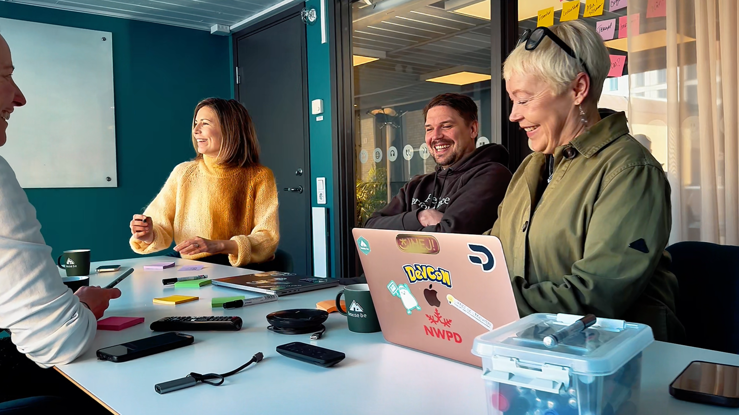Fyra personer som skrattar och samtalar i ett mötesrum. Det finns kaffe muggar, smartphones och en laptop med klisterlappar och stickers. Rummet har ett fönster med post-it lappar på. Träväggar och en whiteboard."
