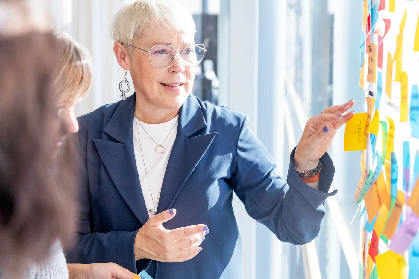 En kvinna med kort blont hår, glasögon och smycken pratar med andra personer framför en vägg med många färgglada post-it-lappar.