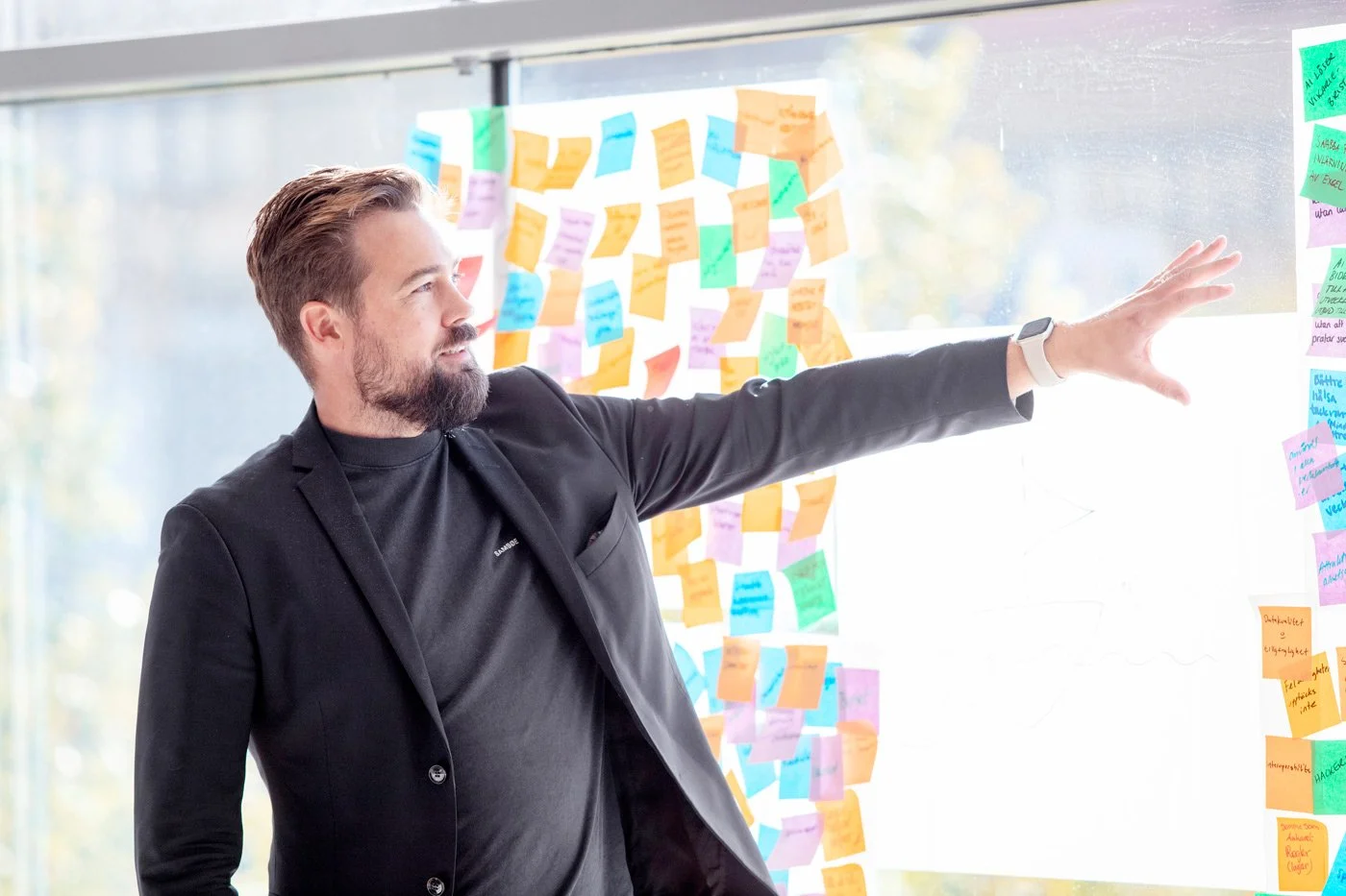 Man i svart jacka och svart tröja som pekar på en whiteboard täckt av färgglada post-it-lappar.