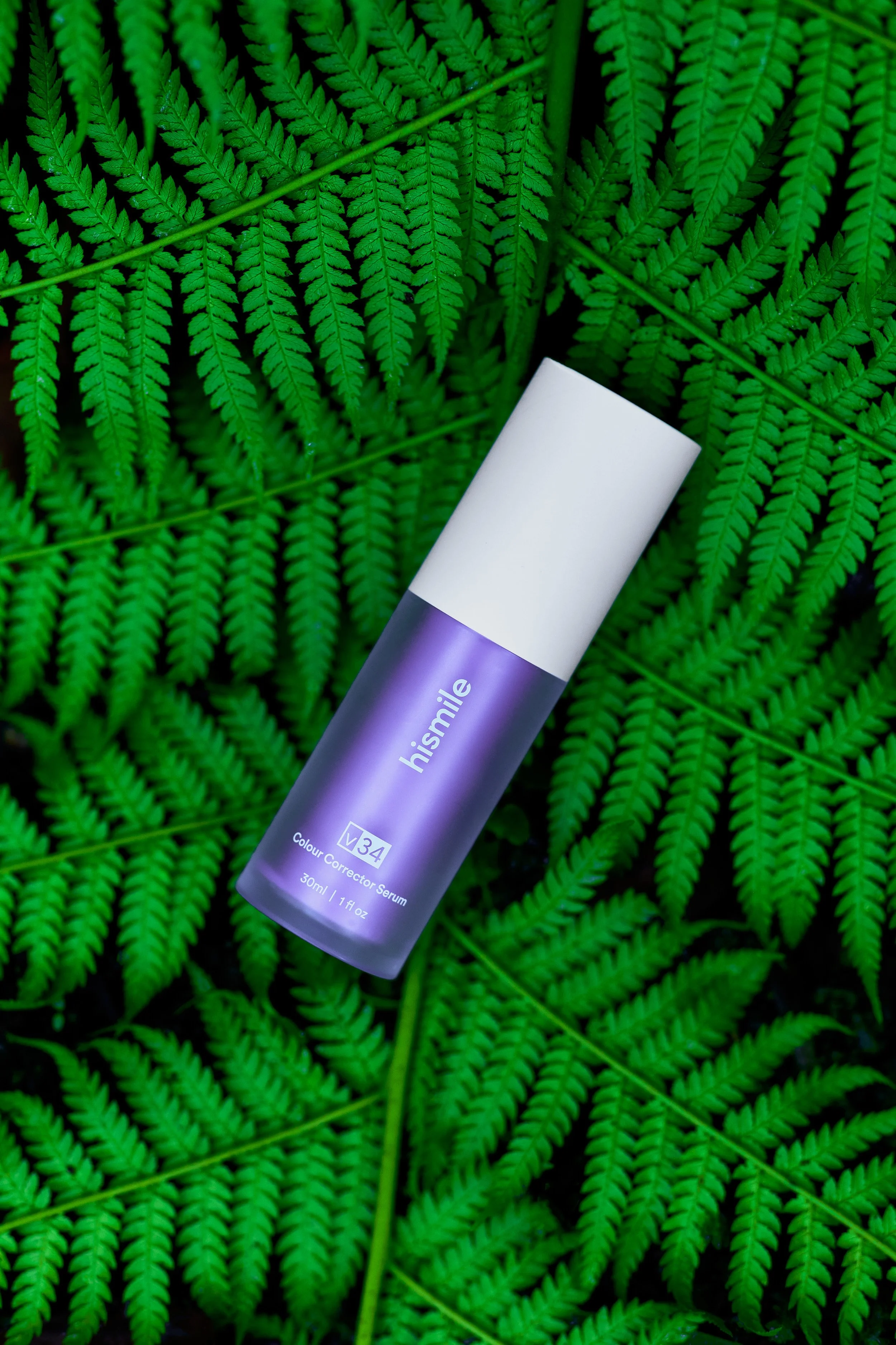 A purple and white bottle of hismile colour corrector serum lying on green fern leaves.