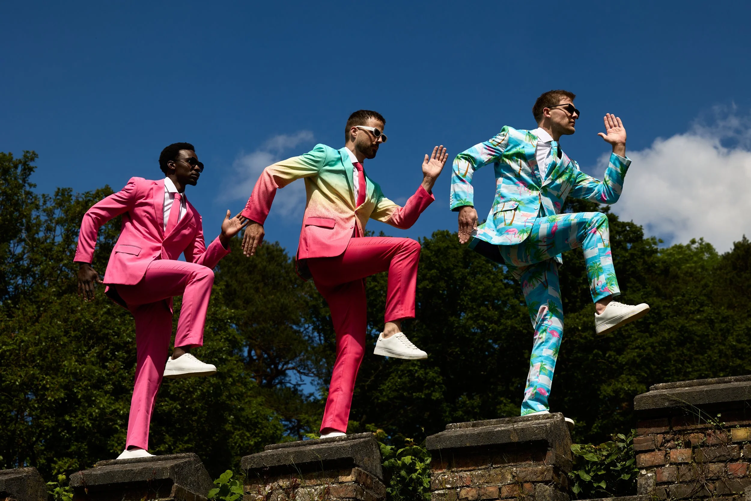 Four men dressed in colorful suits (pink, rainbow, and tropical pattern) walking up a brick ledge, each raising one hand in a running gesture, outdoors with trees and blue sky in the background.