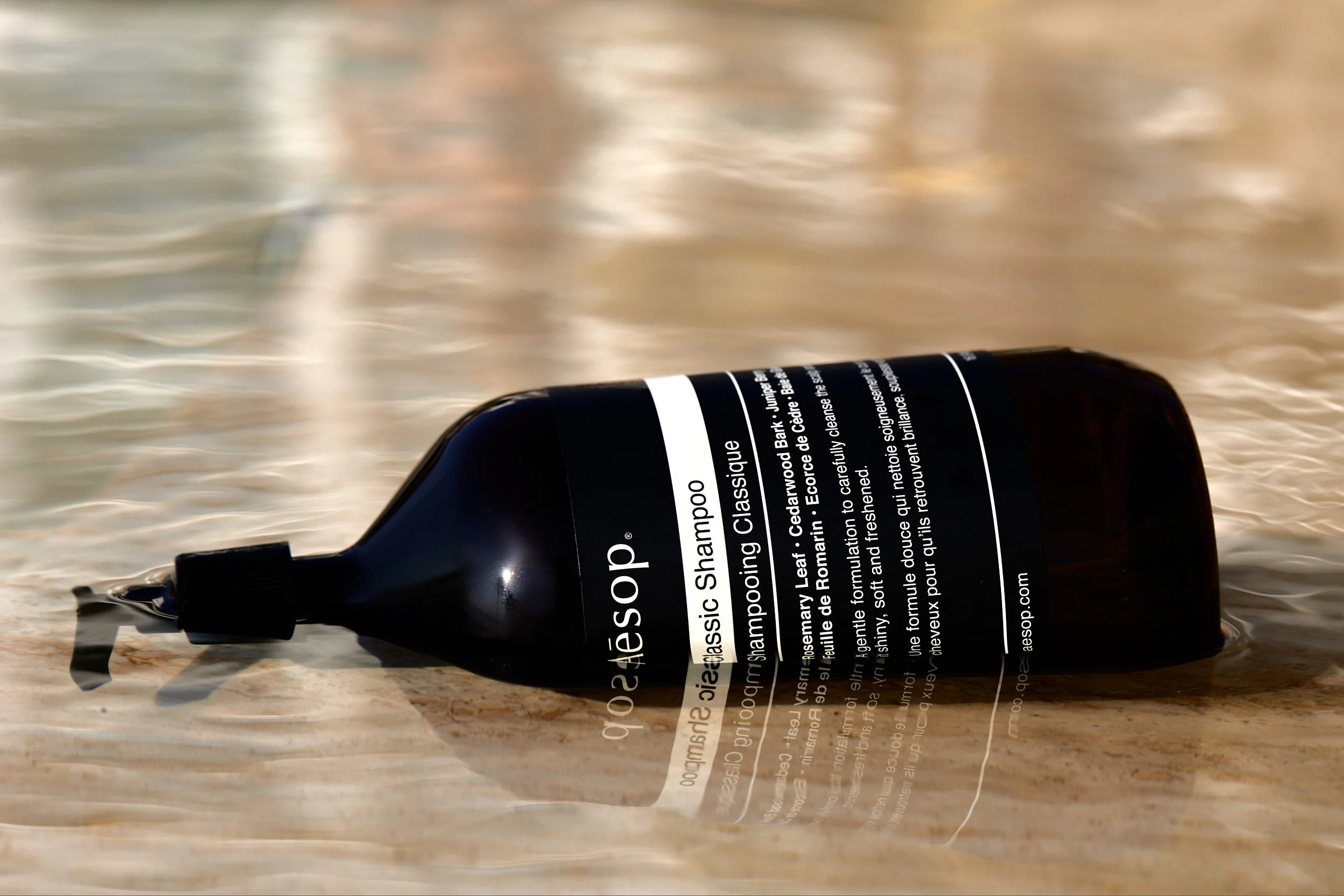 A black bottle of Aesop Classic Shampoo with a pump dispenser lying on a wooden surface near water.