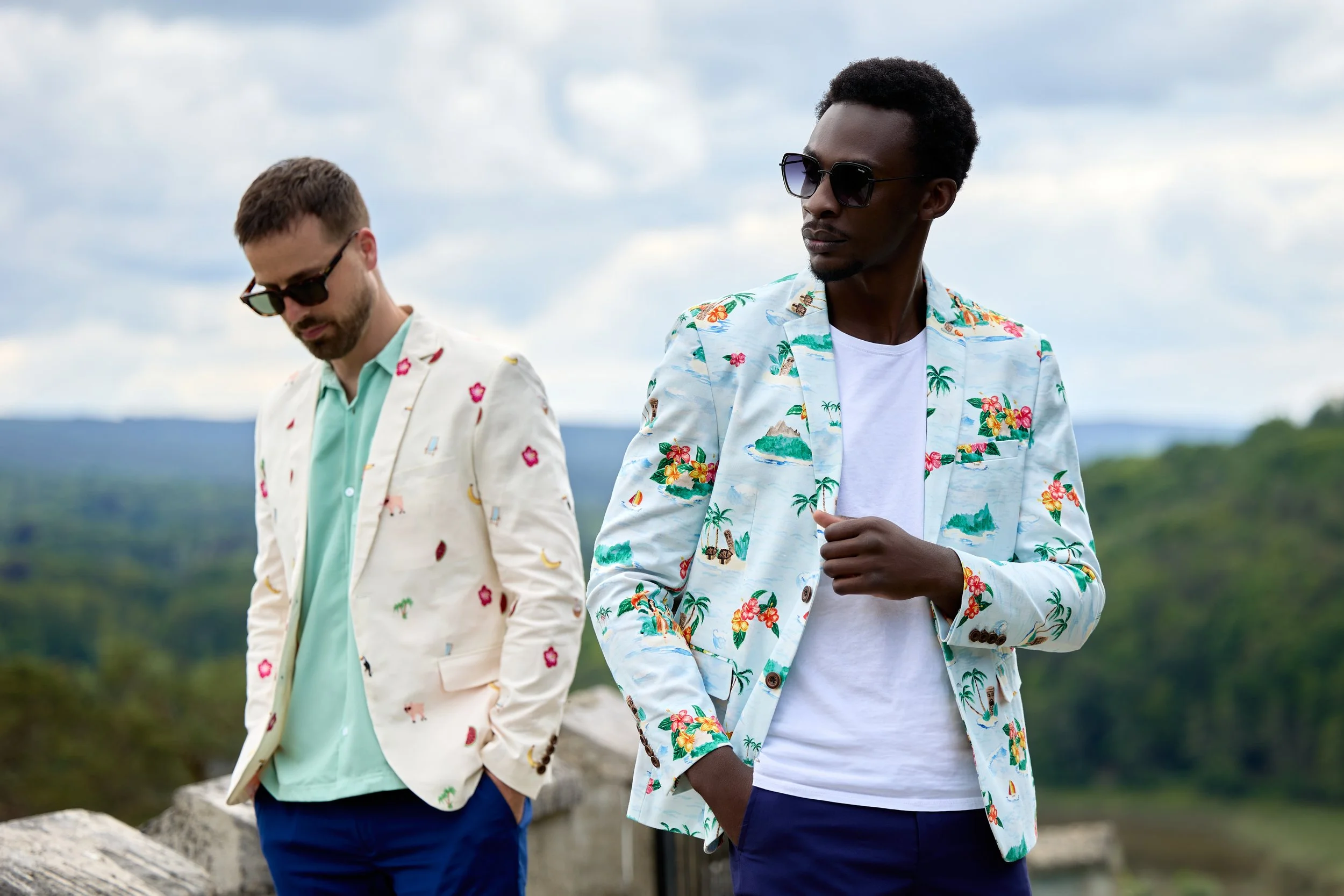 Two men standing outdoors in a scenic natural setting, wearing colorful tropical patterned blazers and sunglasses.