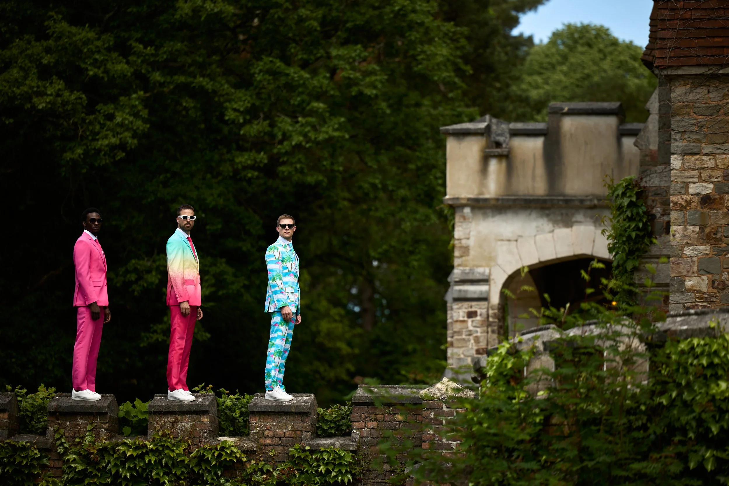 Three men standing on brick columns outdoors, dressed in colorful suits, with a background of green trees and old stone architecture.