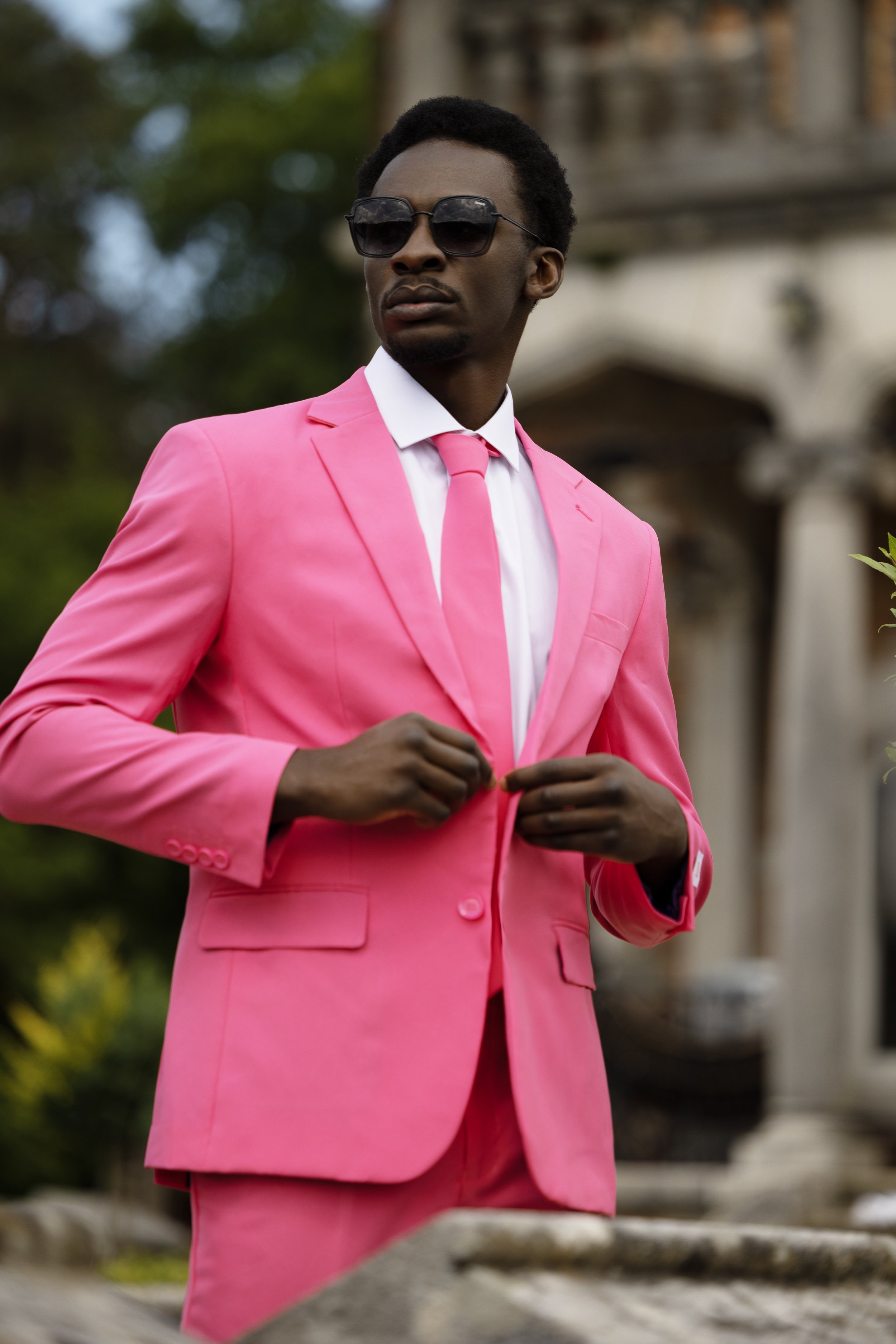A man dressed in a pink suit, white shirt, and pink tie, wearing sunglasses. He is buttoning his suit jacket outdoors.