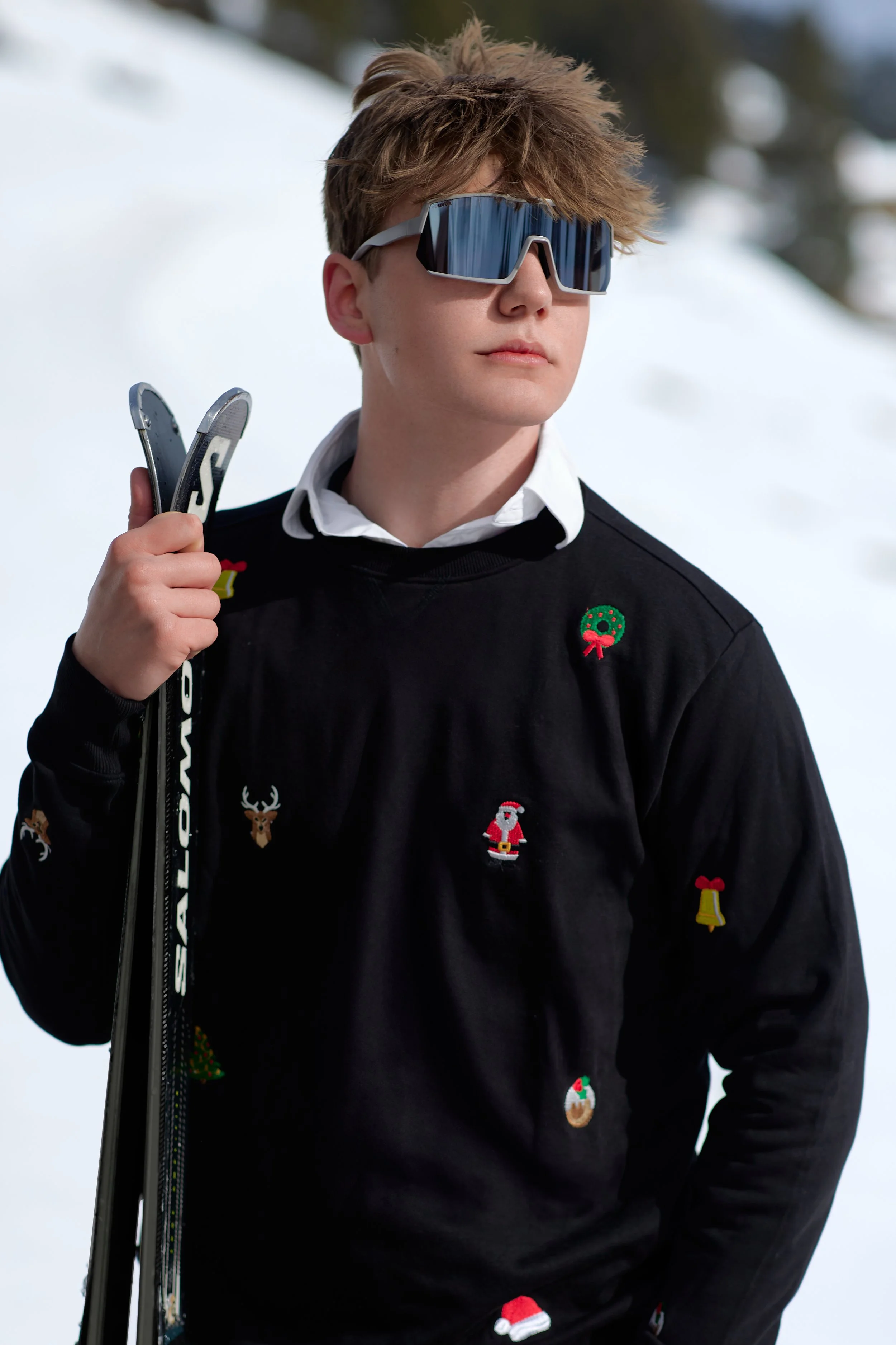 A young man with messy brown hair wearing reflective ski goggles, a black sweater with holiday-themed patches like a Santa, reindeer, and Christmas decorations, holding ski poles in a snowy outdoor setting.