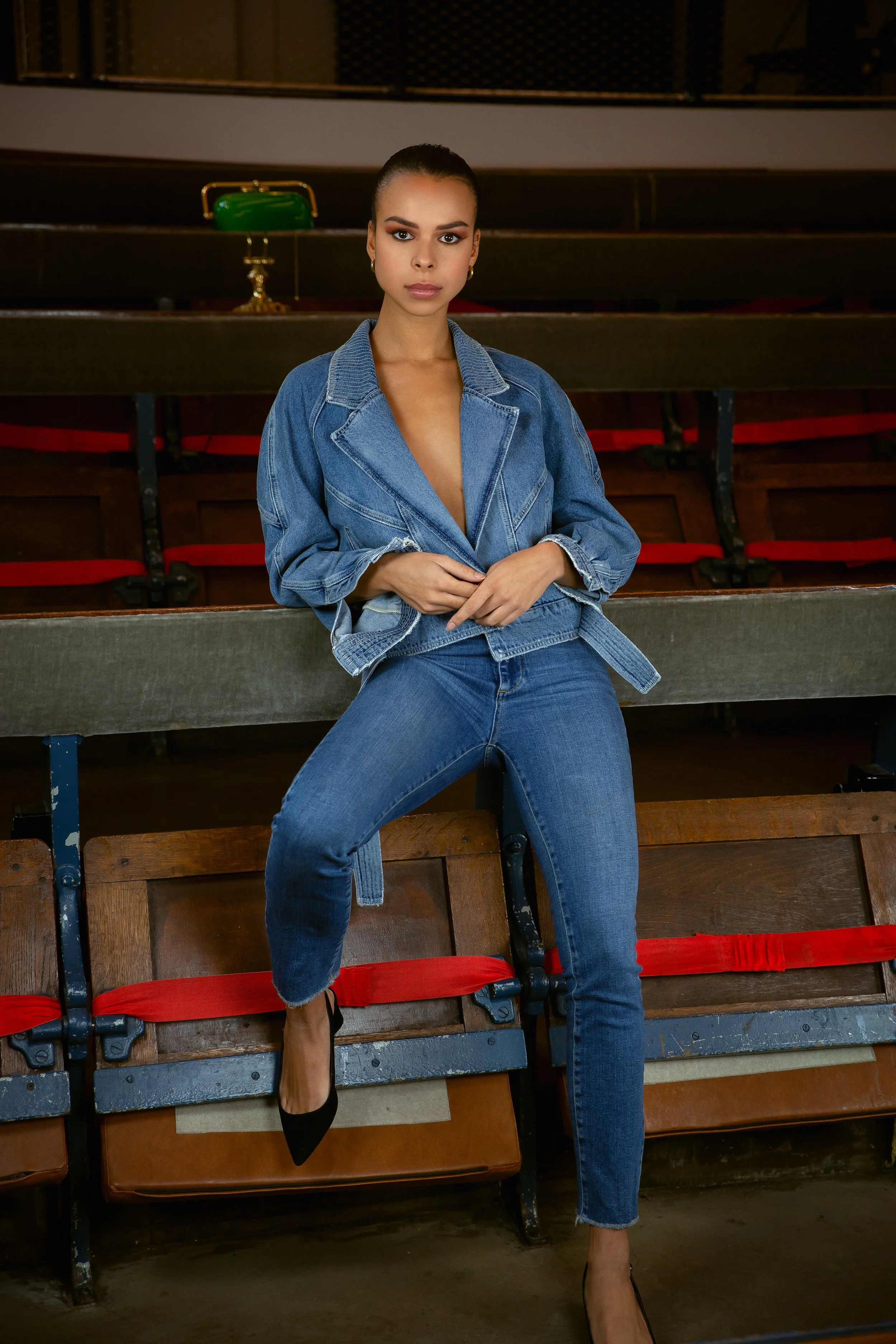 A woman wearing a denim jacket and jeans sitting on a wooden bench in an empty auditorium with red and black seats.