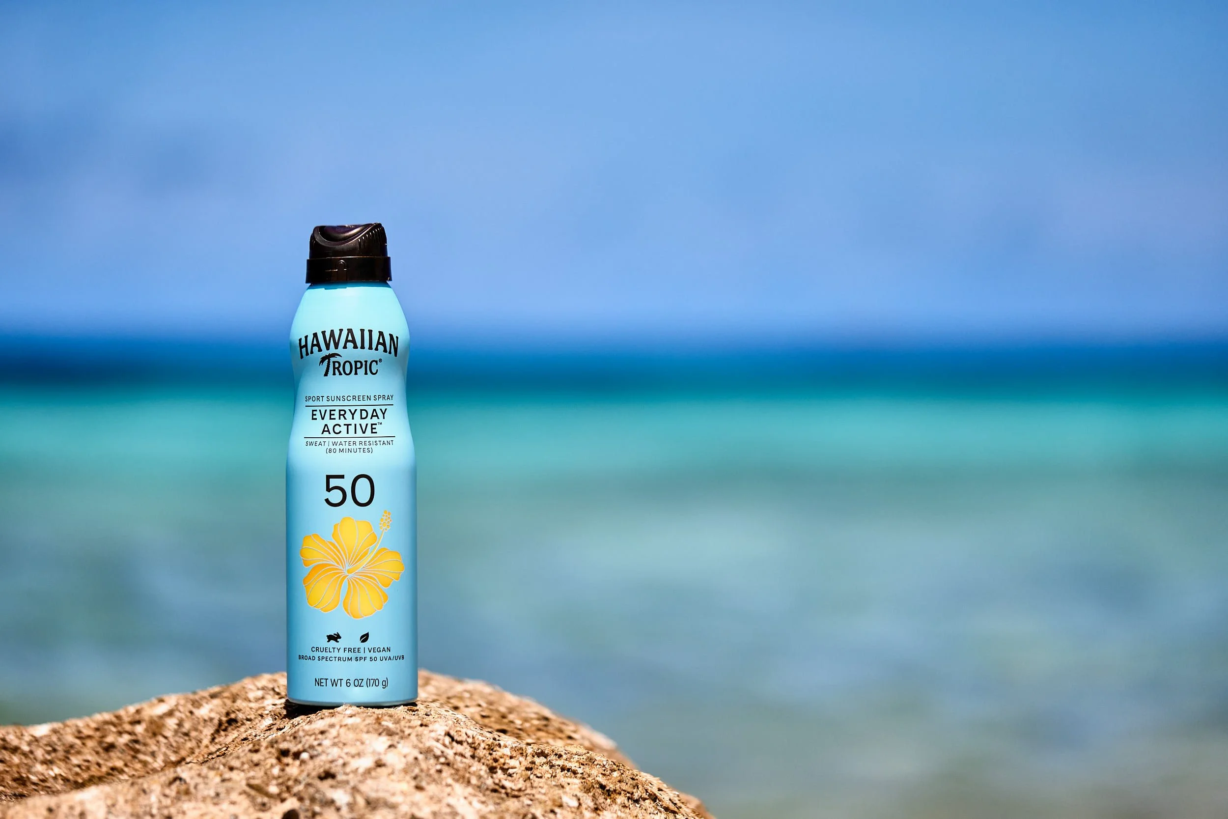 A bottle of Hawaiian Tropic sunscreen spray with SPF 50 on a rock at the beach, with ocean and sky in the background.