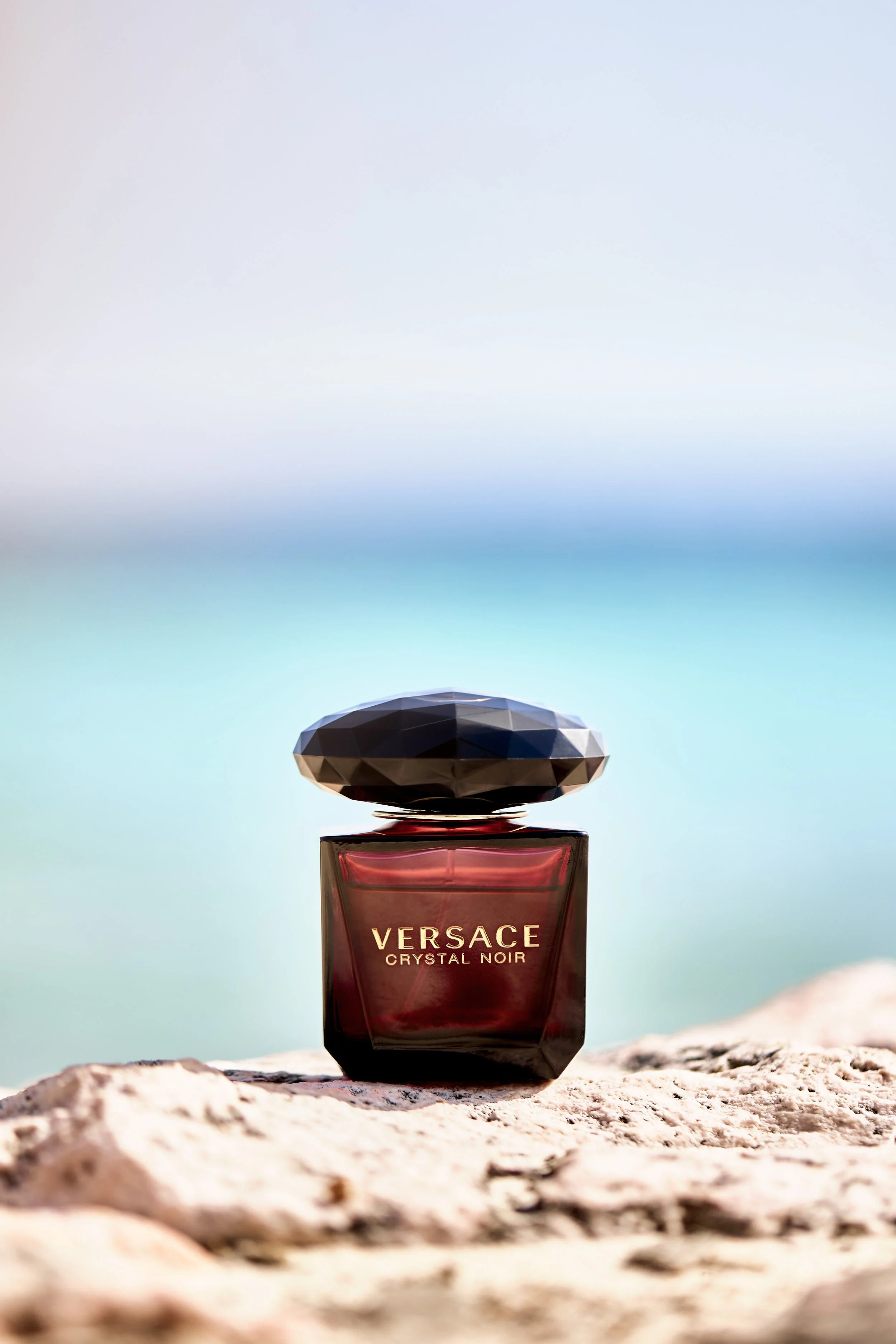 A bottle of Versace Crystal Noir perfume placed on a rocky surface with a blurred beach and sky background.