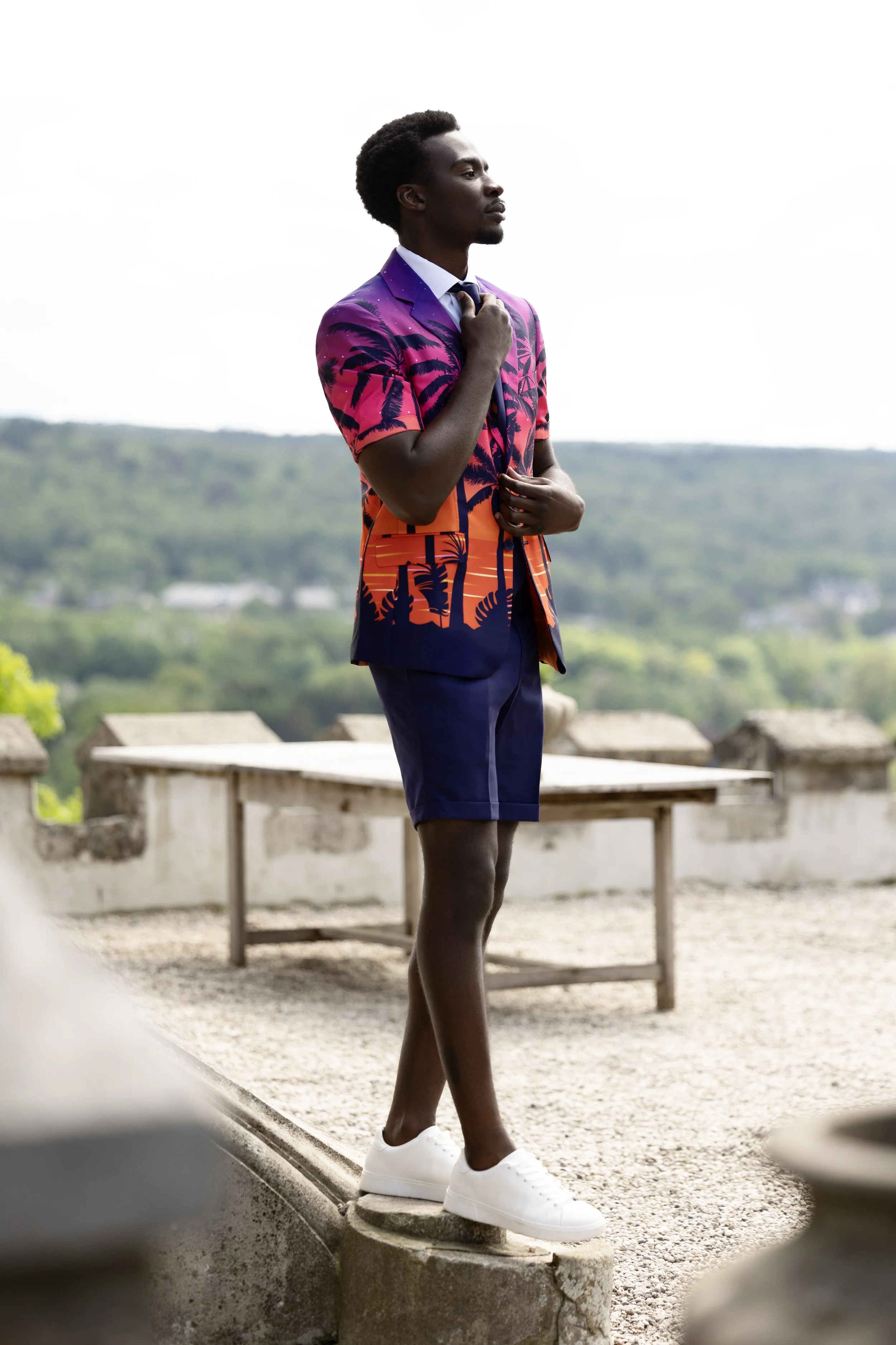 A young man standing outdoors on a terrace, wearing a colorful short-sleeved blazer with a tropical print, dark shorts, and white sneakers, with a scenic landscape in the background.
