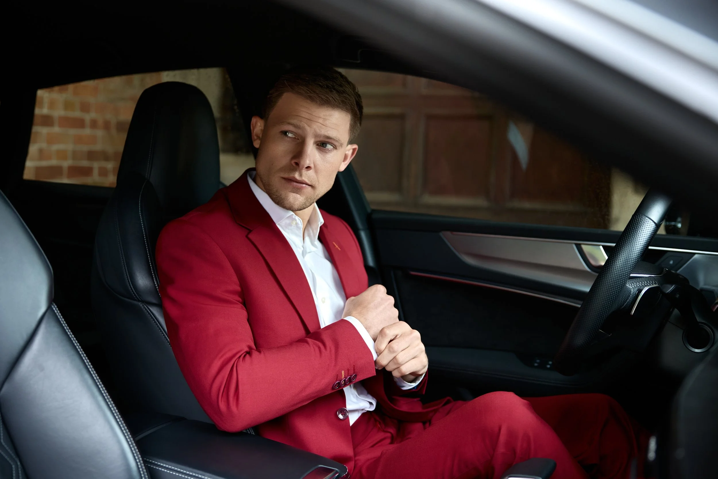 A man in a red suit sitting inside a car, looking to his left.