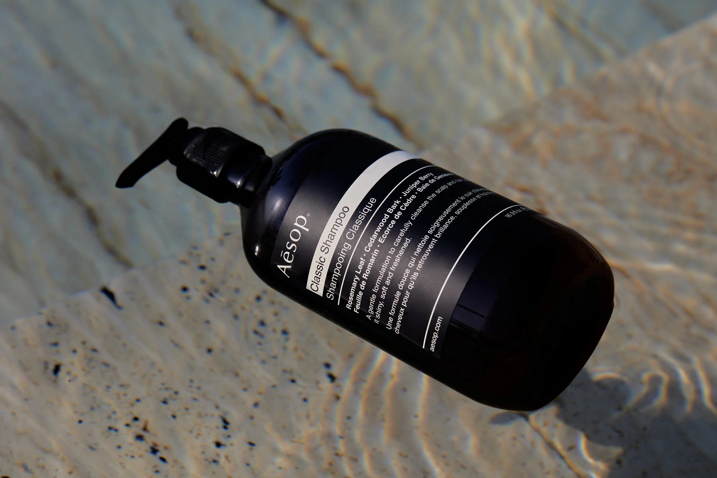 A black bottle of Aésop Classic Shampoo lying on wet sand near water, with a pump dispenser on top.