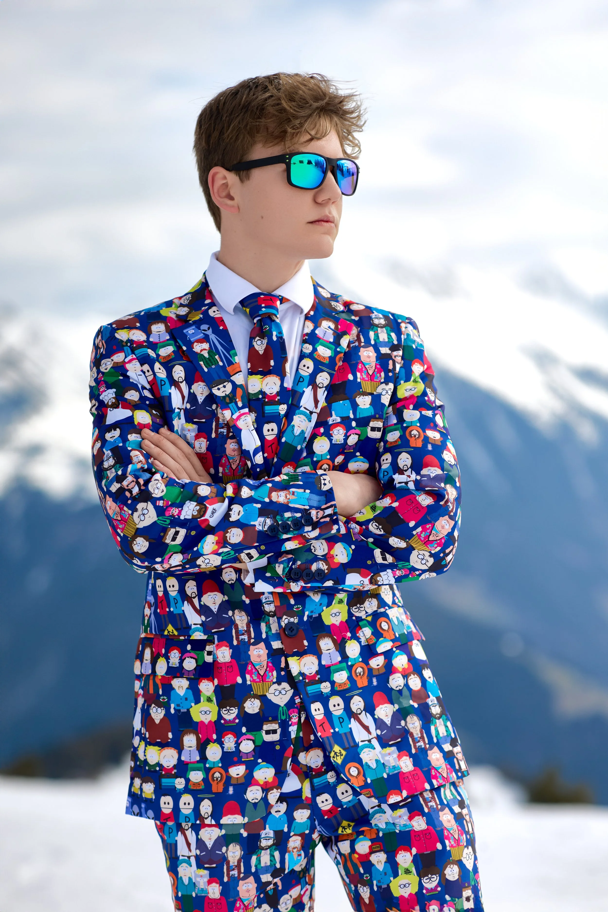 A young person dressed in a colorful suit featuring cartoon characters, standing outdoors in a snowy mountain landscape with arms crossed and wearing reflective blue sunglasses.