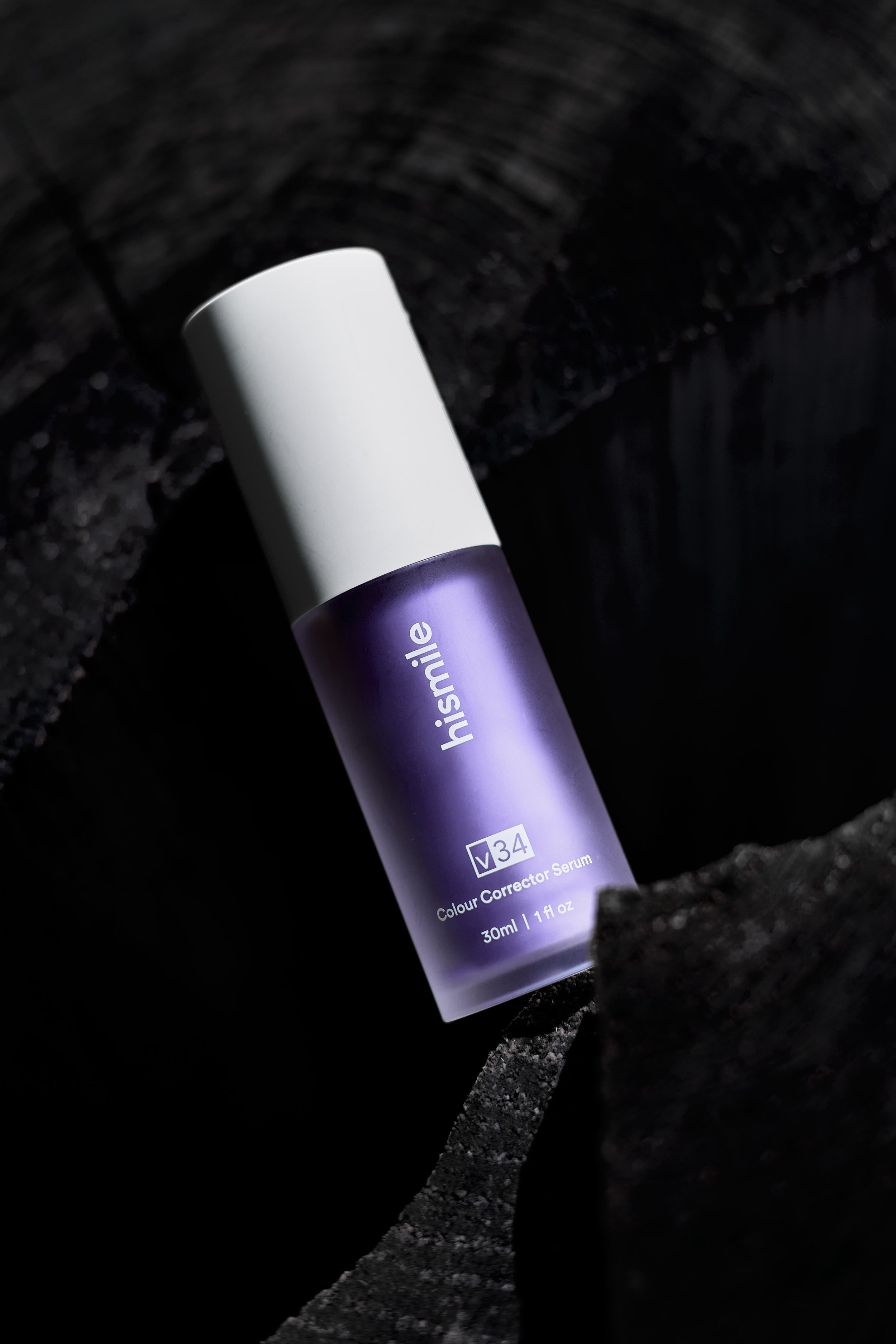 A small purple bottle of Iismile colour corrector serum with a white cap, placed on a dark textured surface.