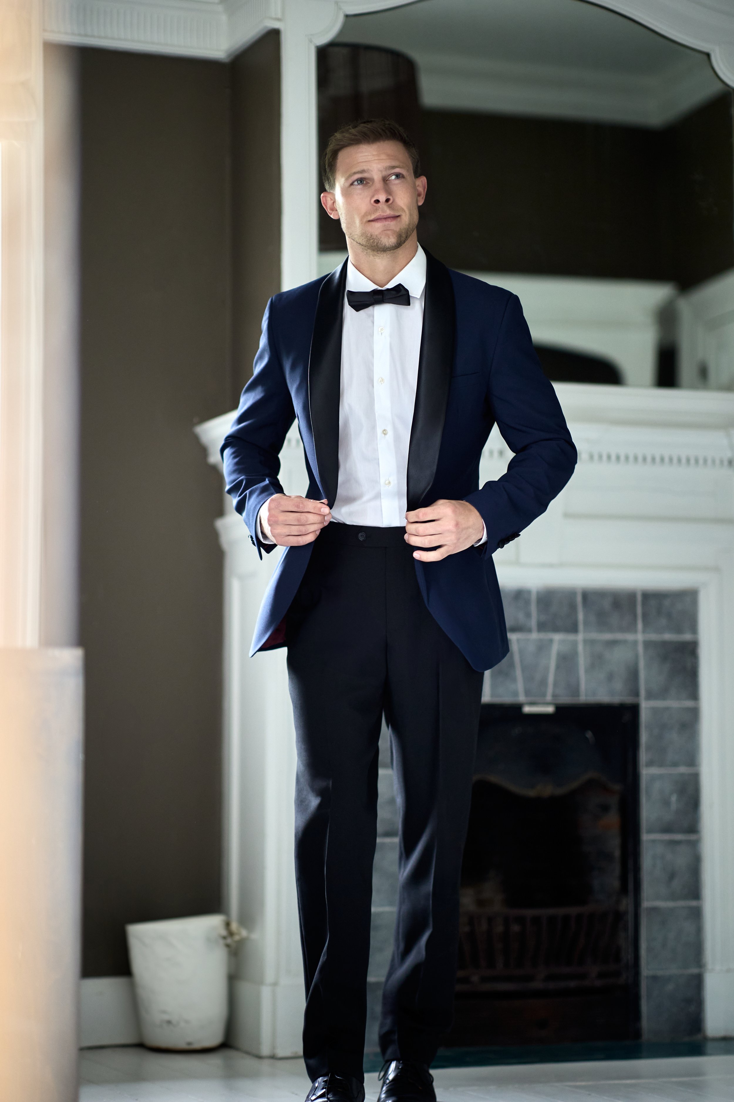A man in a navy tuxedo with black satin lapels, white dress shirt, and black bow tie adjusts his jacket in front of a fireplace.